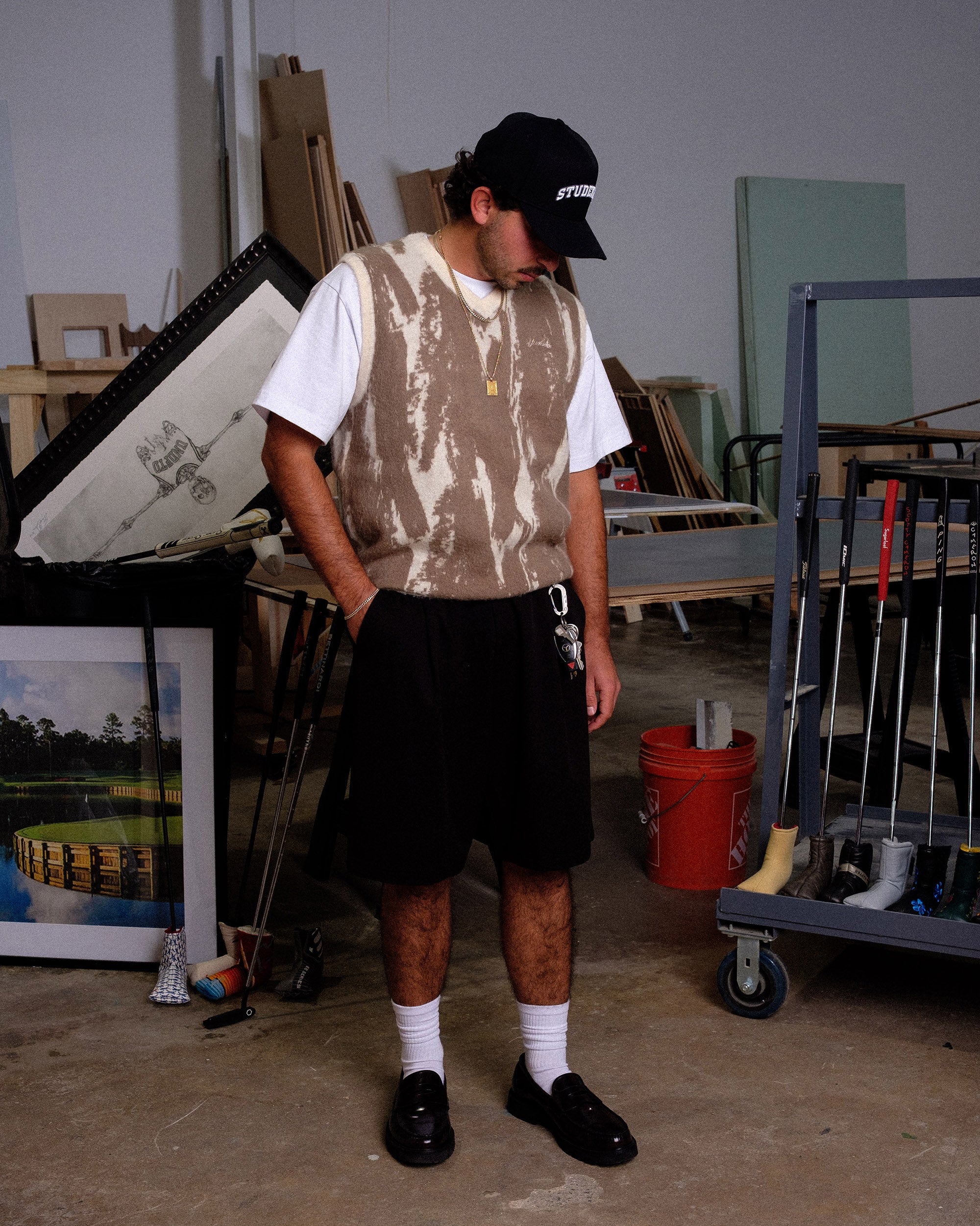 Person wearing a camouflage vest and black cap in an indoor setting with various items around