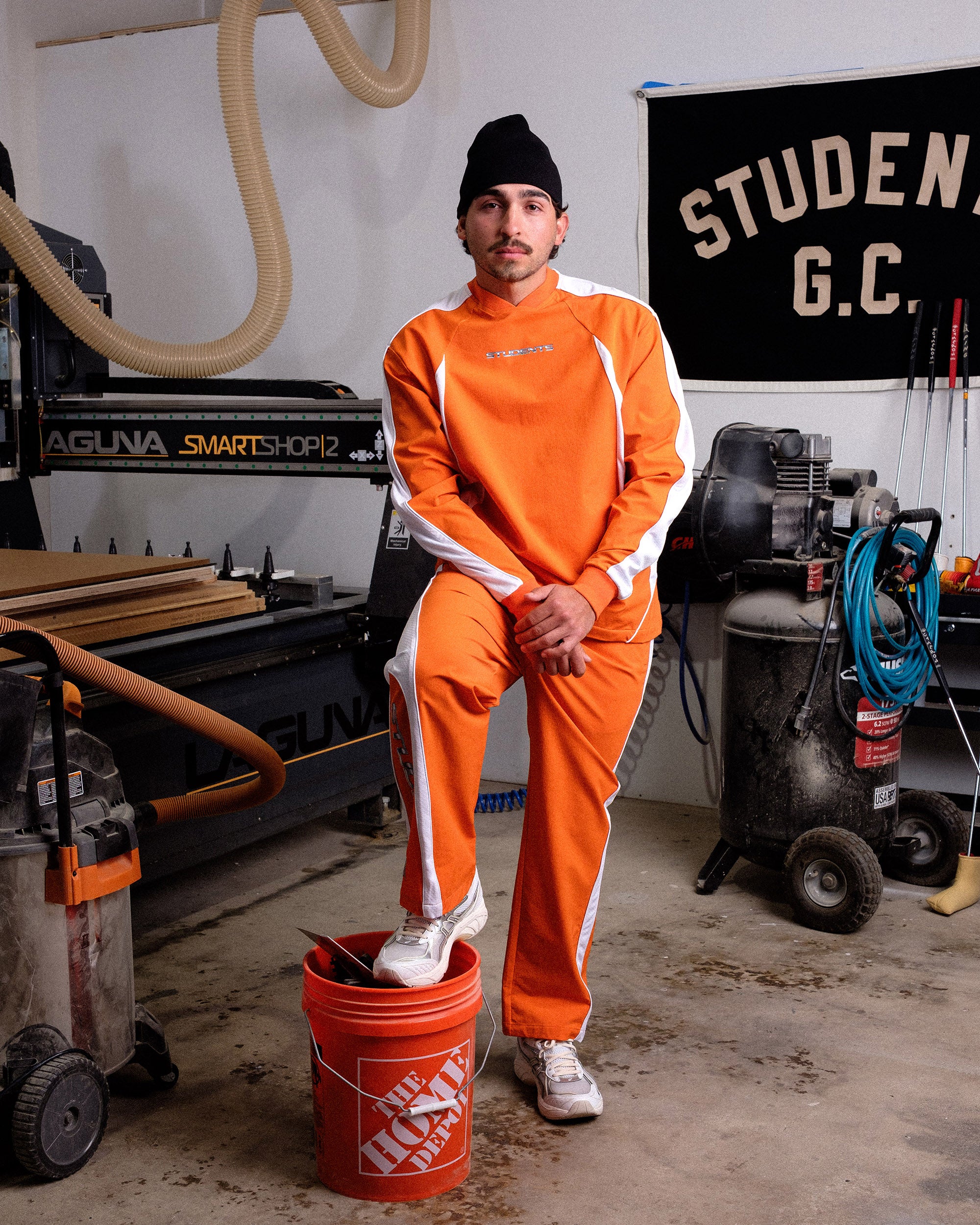 Person in orange jumpsuit standing next to a red bucket in a workshop with 'STUDENT G.C.' sign.