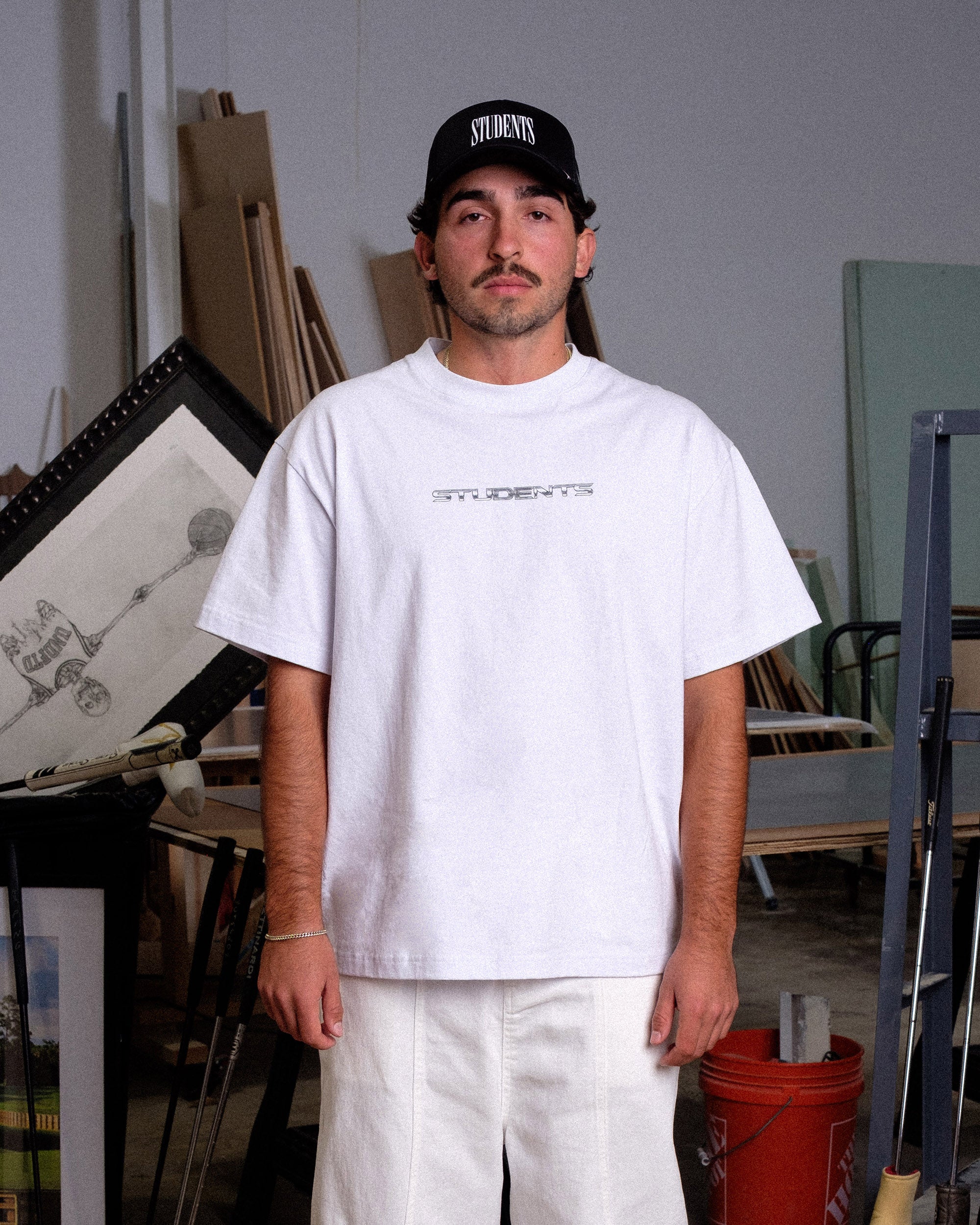Man wearing a white t-shirt and black cap in an indoor setting with art supplies.