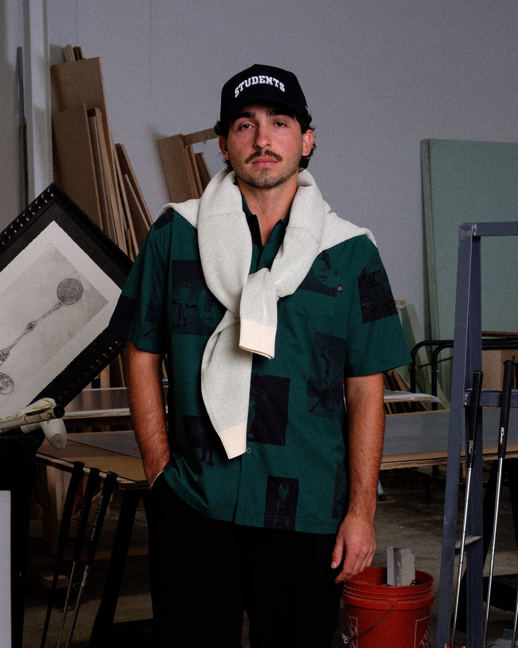 Man wearing a cap and scarf in an indoor setting with art supplies and furniture.