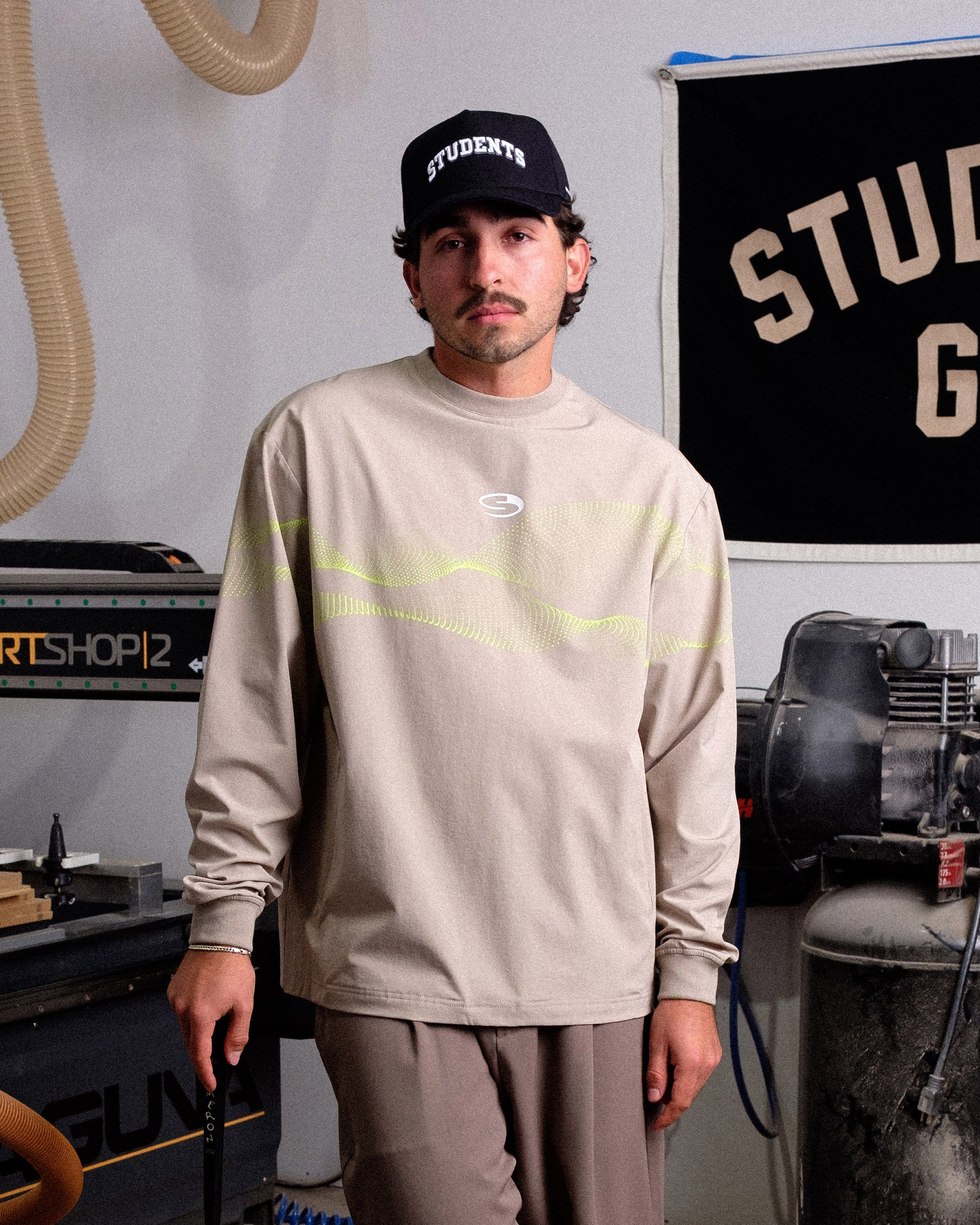 Man wearing a beige long-sleeve shirt with neon accents and a black cap in a workshop setting.