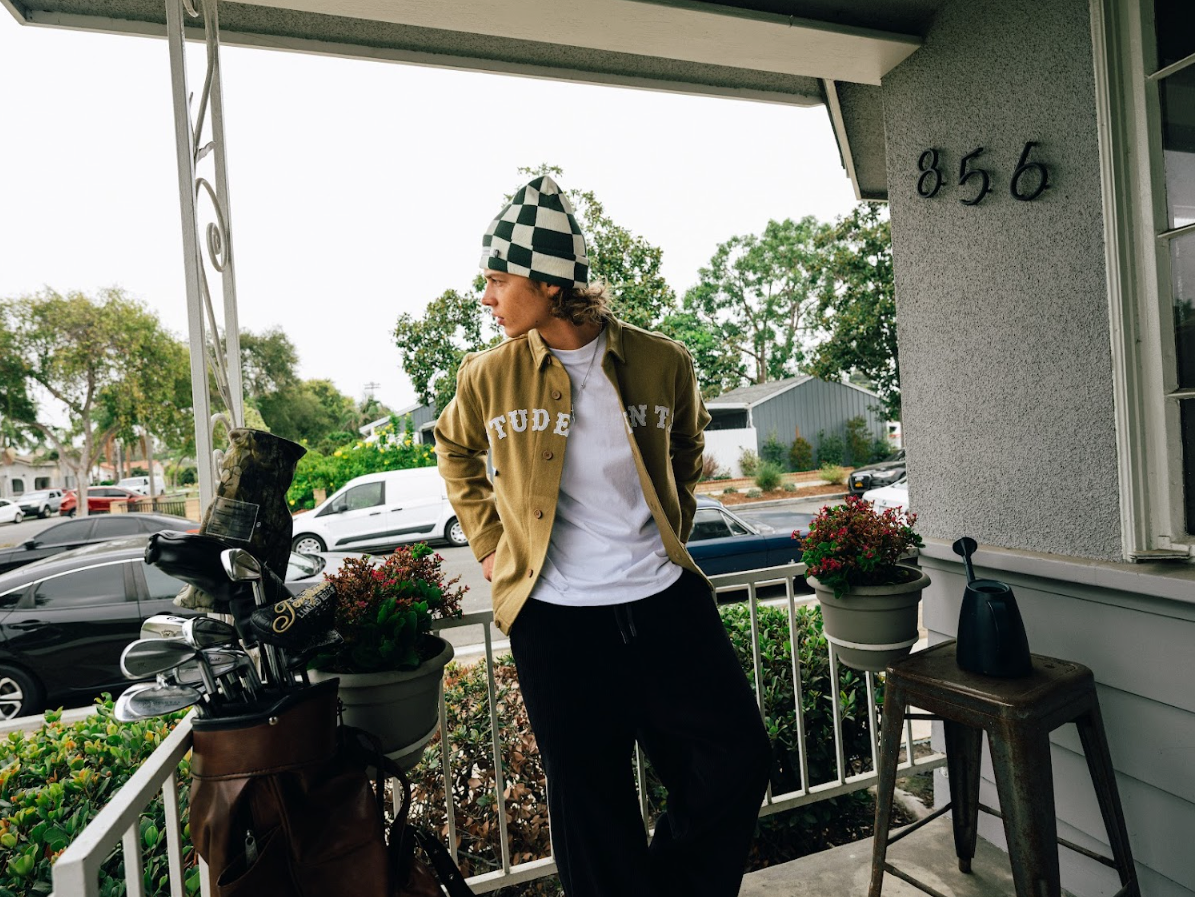 Person standing on a porch wearing a checkered beanie and a yellow jacket with text.