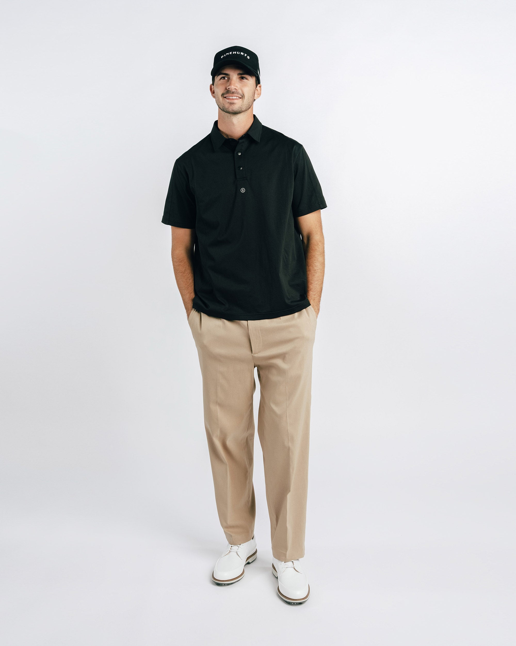 Man wearing a black polo shirt, beige pants, and white shoes on a white background