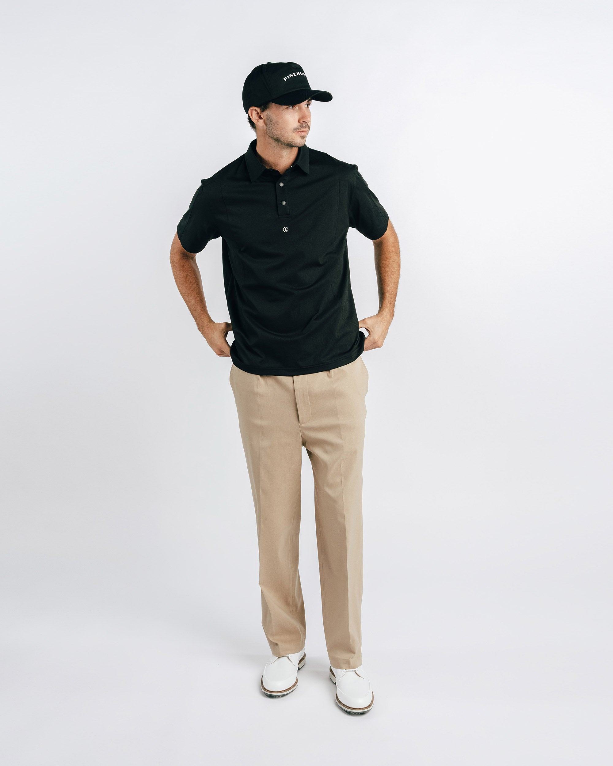 Man wearing a black polo shirt, beige pants, and white shoes on a white background
