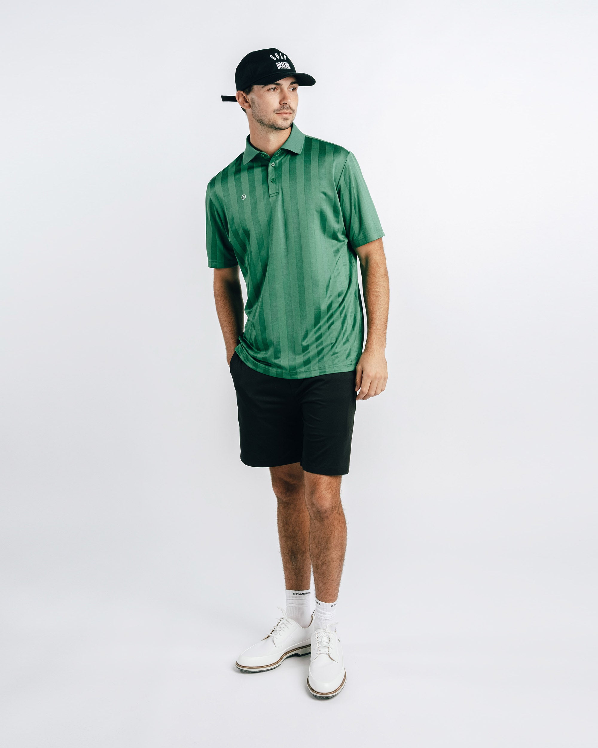 Man wearing a green polo shirt, black shorts, white shoes, and a black cap on a white background