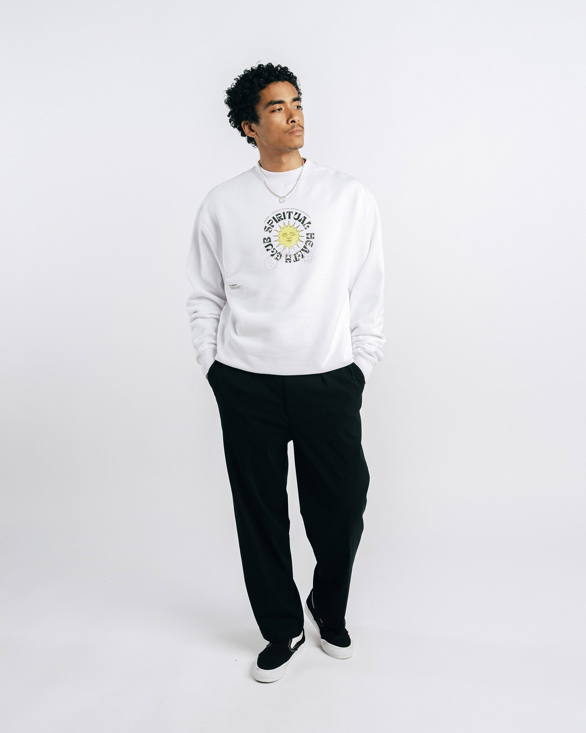 Person wearing a white sweater with a logo and text, black pants, and white shoes on a white background