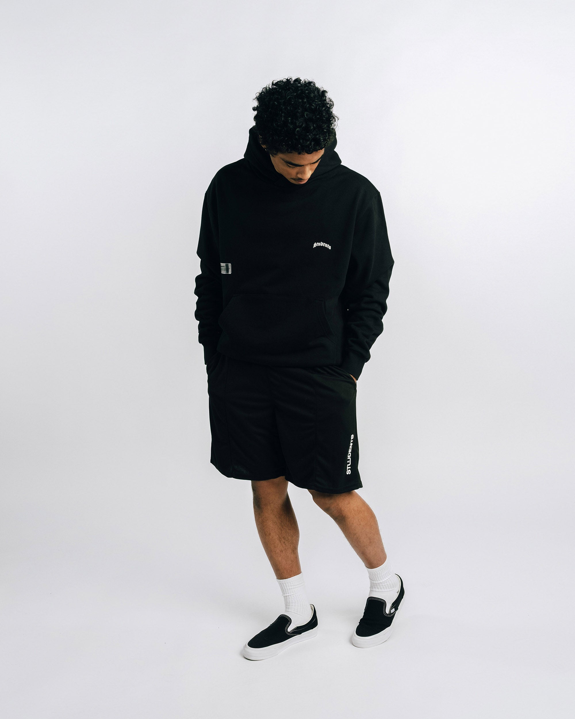 Person wearing a black hoodie and shorts set with white socks and black shoes on a white background