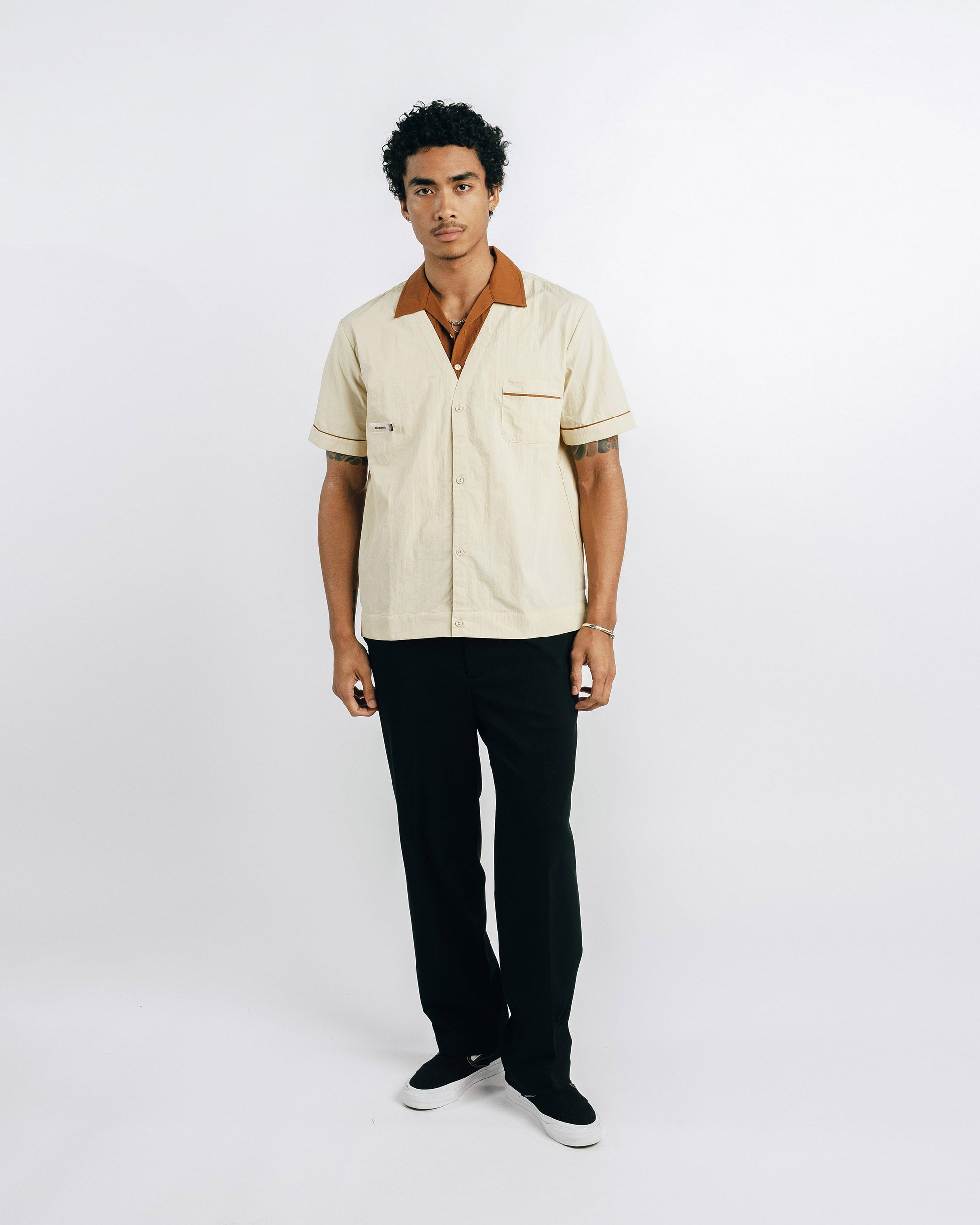 Man wearing a beige shirt with a brown collar and black pants on a white background