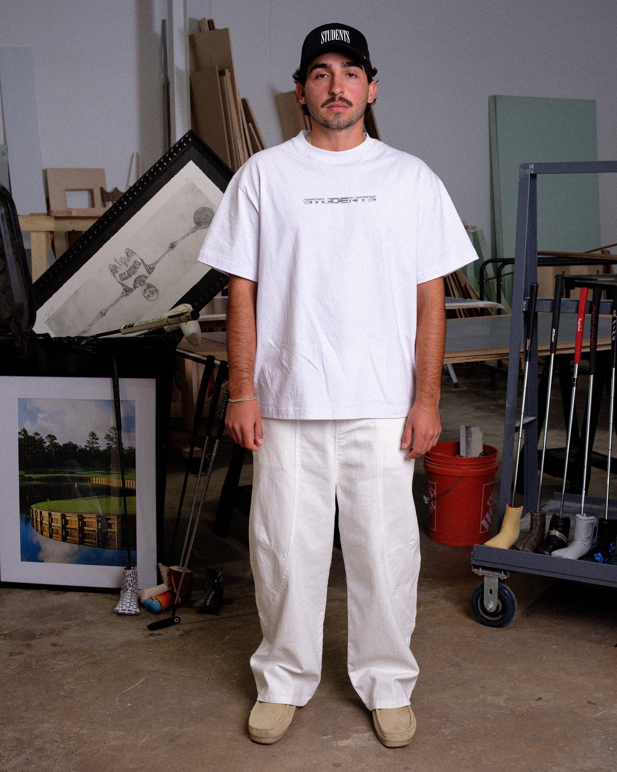 Man wearing a white t-shirt and pants standing in an indoor setting with various items around