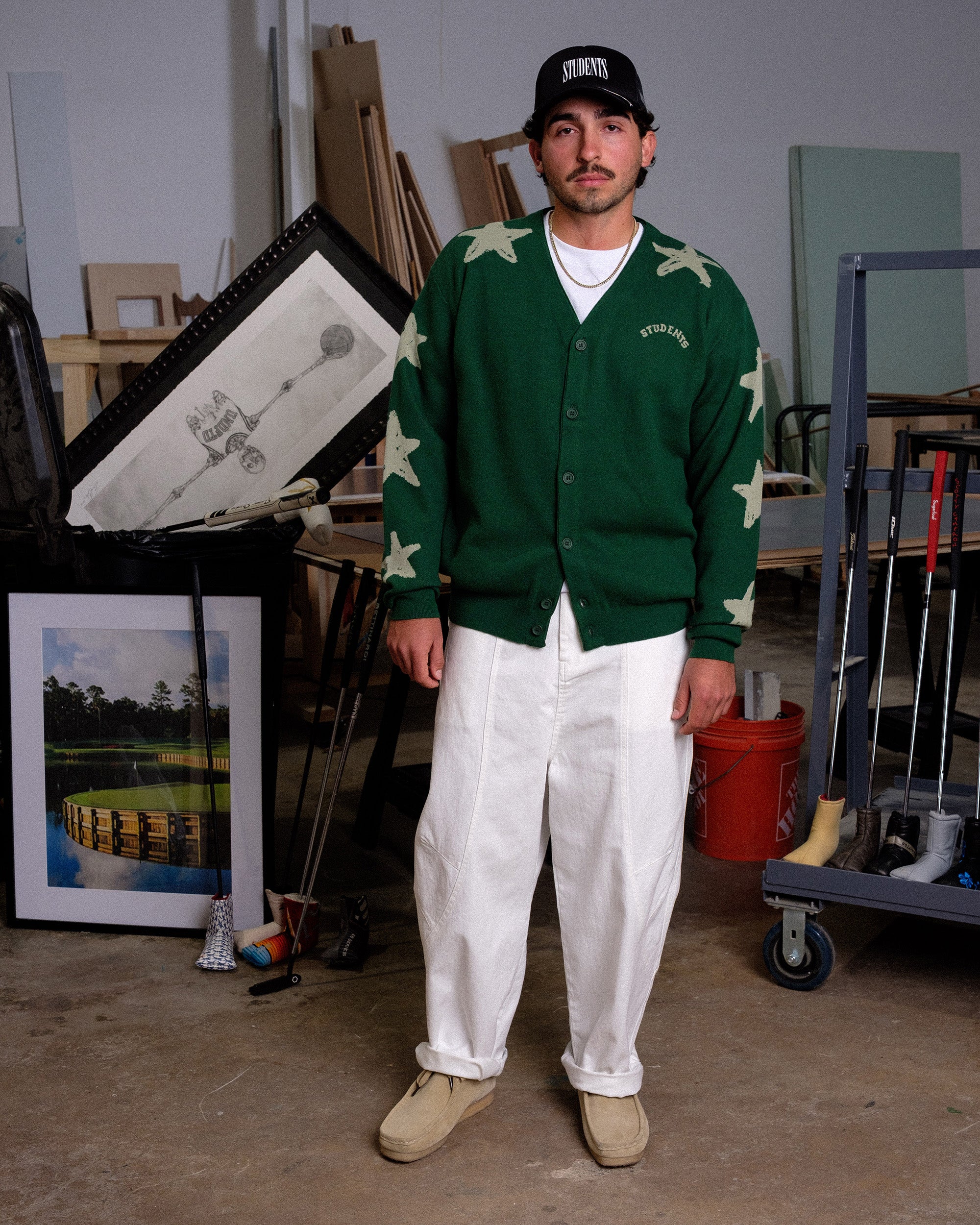 Man wearing a green cardigan with star patterns and white pants standing in an indoor setting.