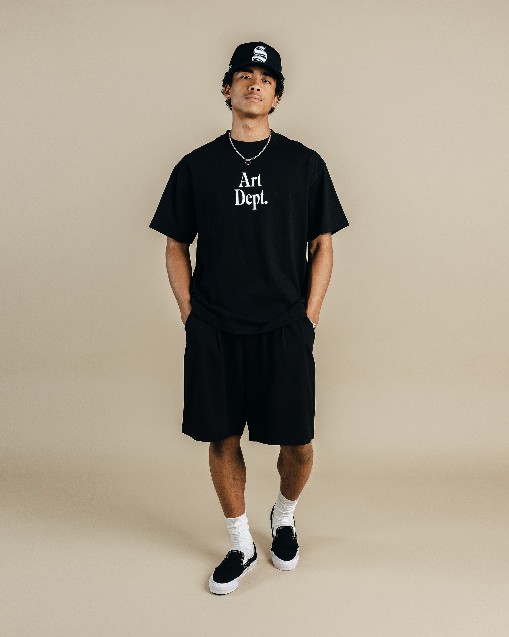 Person wearing a black 'Art Dept.' t-shirt, black shorts, and a black cap on a beige background