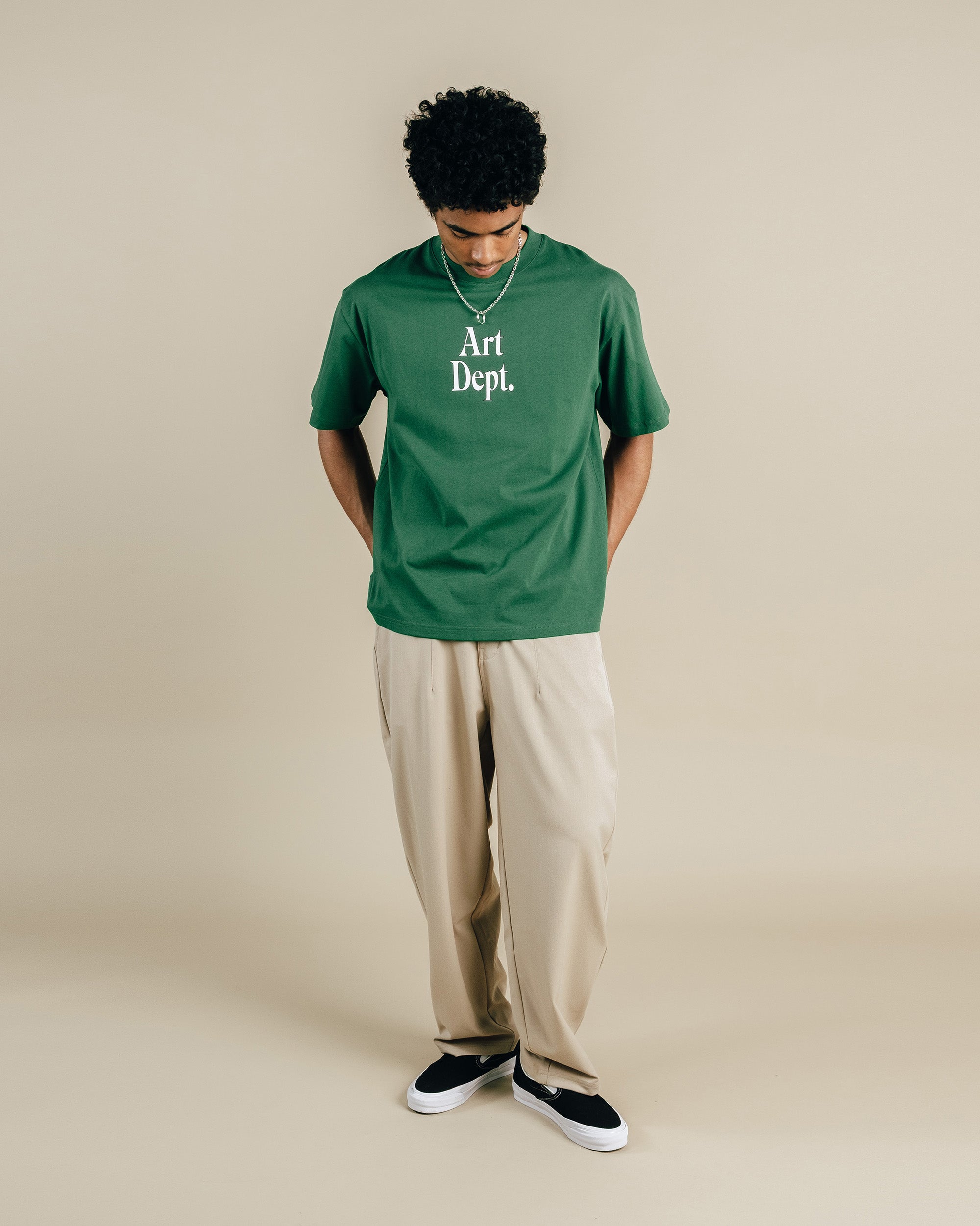 Person wearing a green 'Art Dept.' t-shirt and beige pants on a beige background