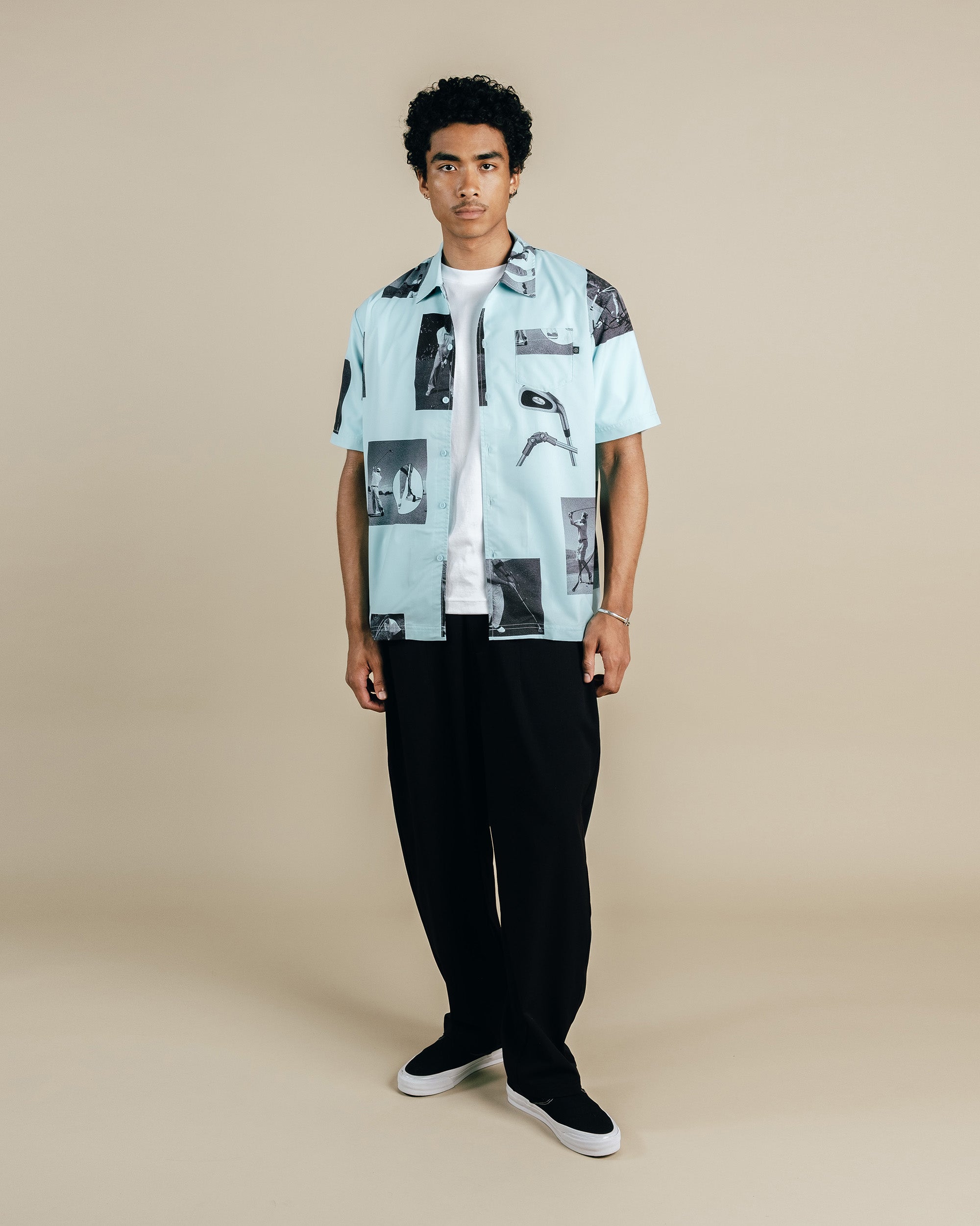 Man wearing a light blue patterned shirt over a white t-shirt with black pants on a beige background