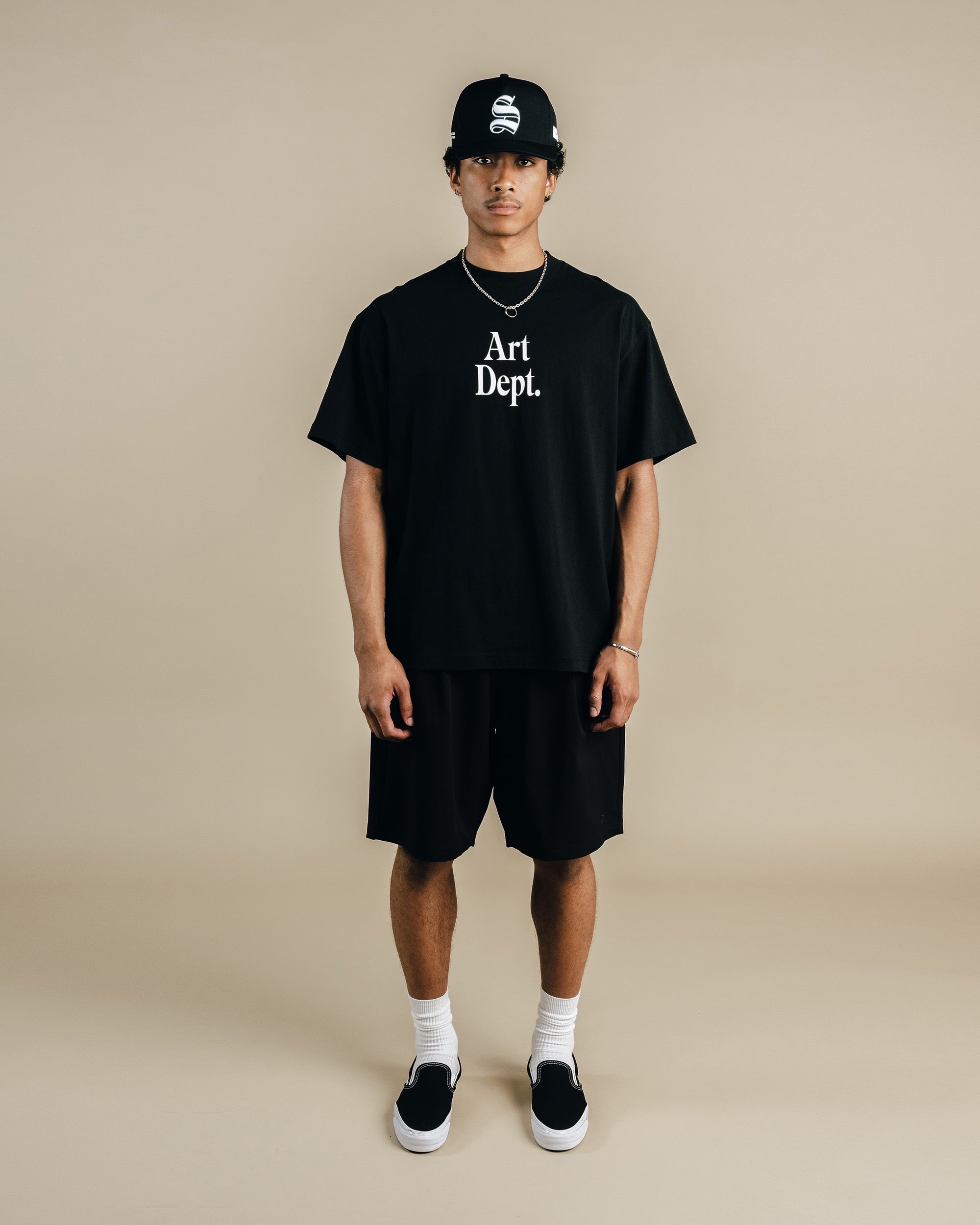Person wearing a black 'Art Dept.' t-shirt, black shorts, and a black cap on a beige background