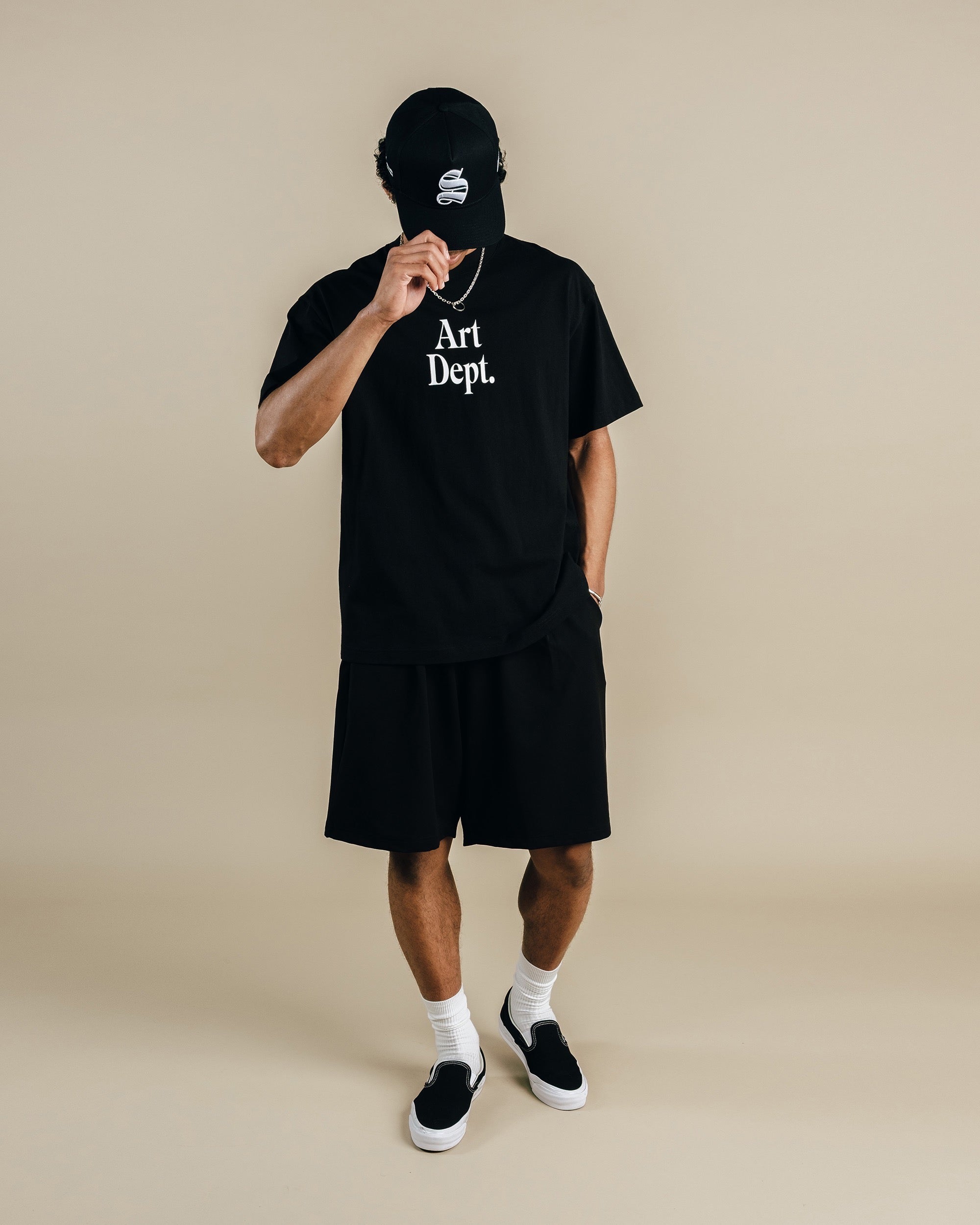 Person wearing a black 'Art Dept.' t-shirt and shorts with a cap on a beige background