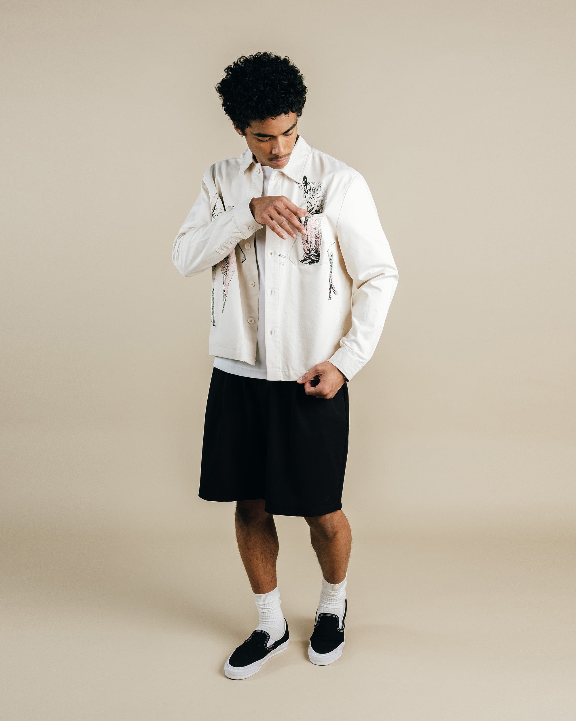 Man wearing a white shirt with black patterns, black shorts, and black shoes on a beige background