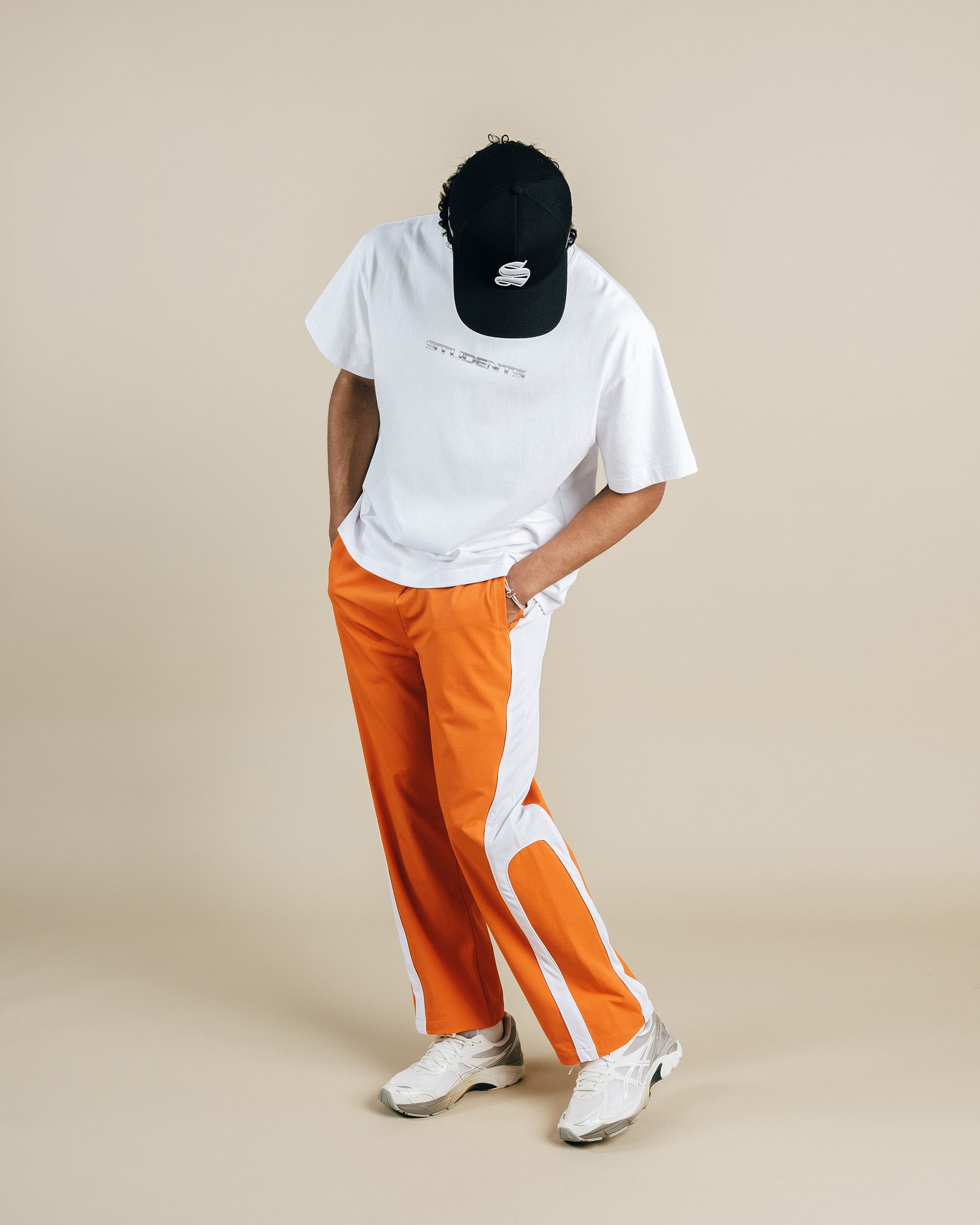 Person wearing a white t-shirt, orange pants with white stripes, and a black cap on a beige background