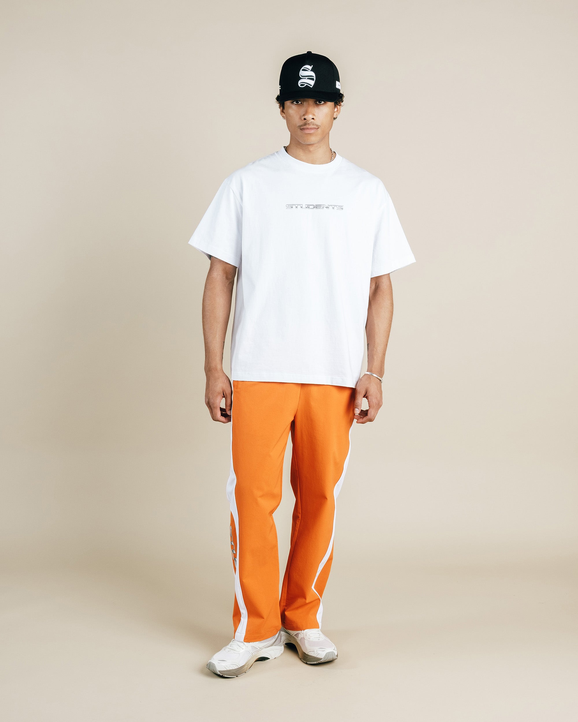 Person wearing a white t-shirt, orange pants, and a black cap on a beige background