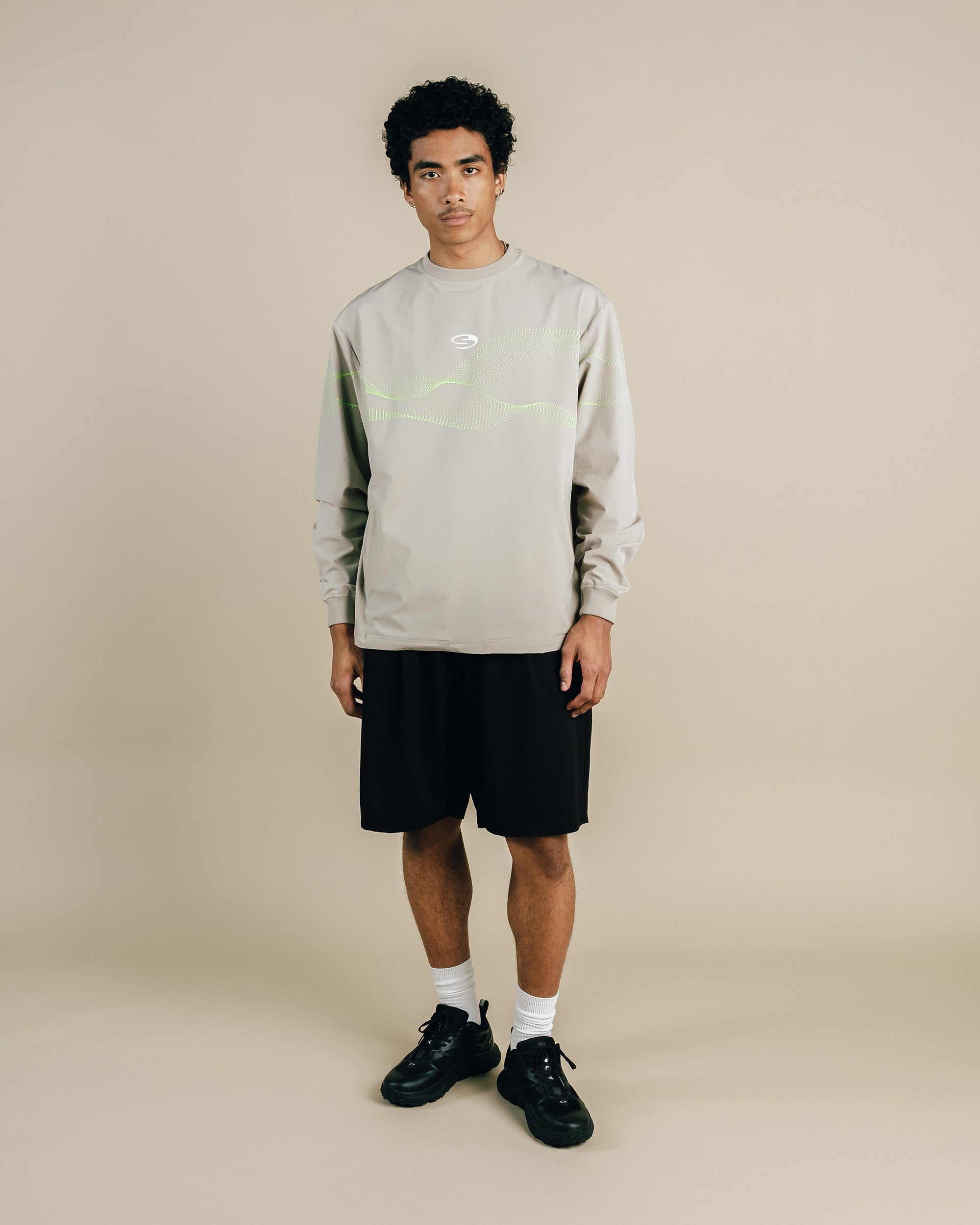 Person wearing a light gray long-sleeve shirt with a logo and black shorts on a beige background