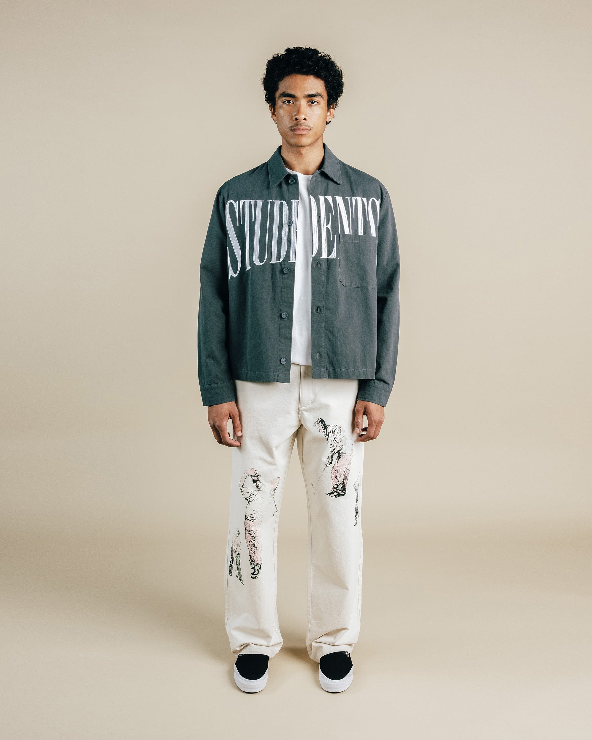 Person wearing a green jacket with 'STUDENTS' text, white shirt, and white pants on a beige background