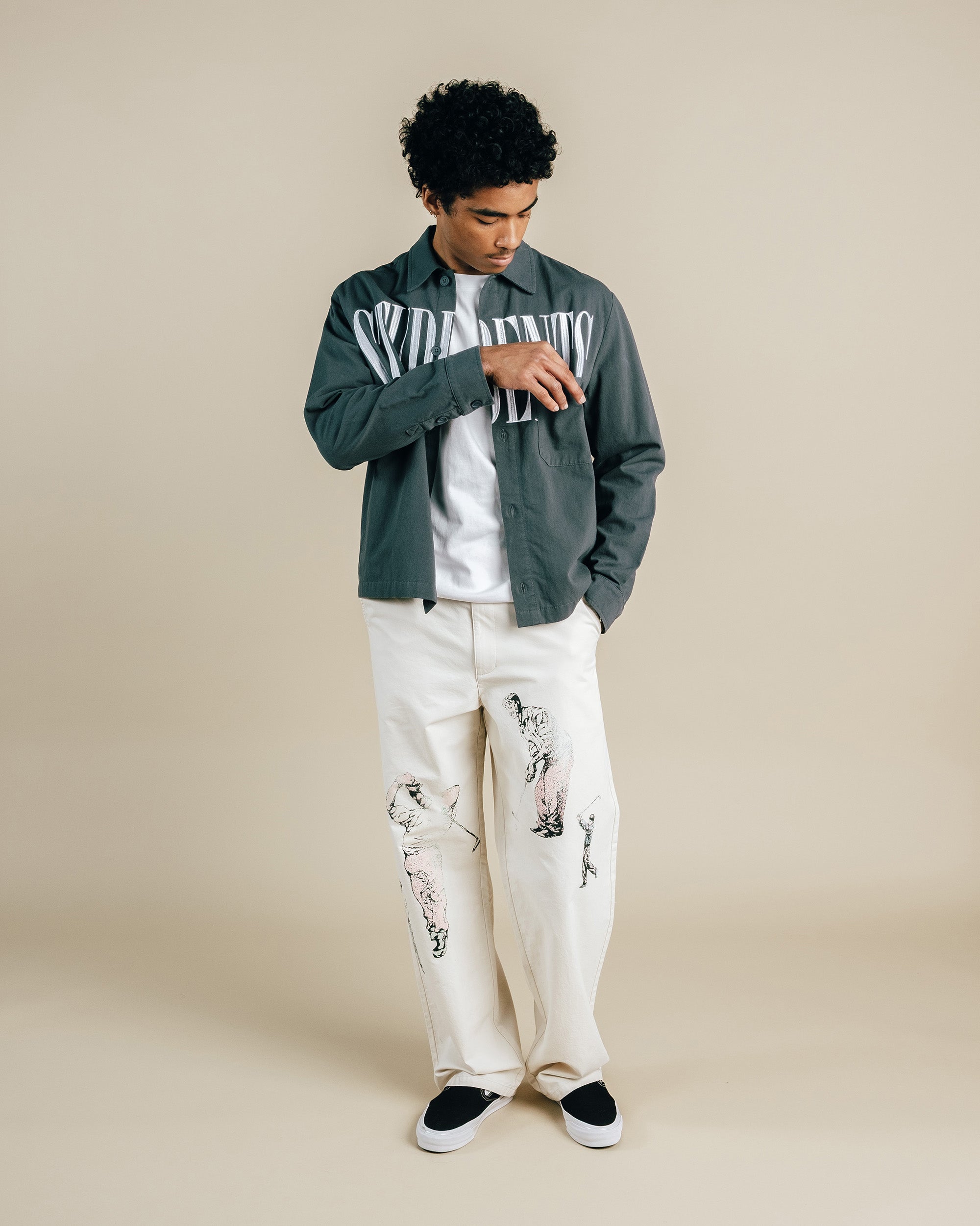 Person wearing a green jacket with text, white shirt, and white pants with black patterns on a beige background