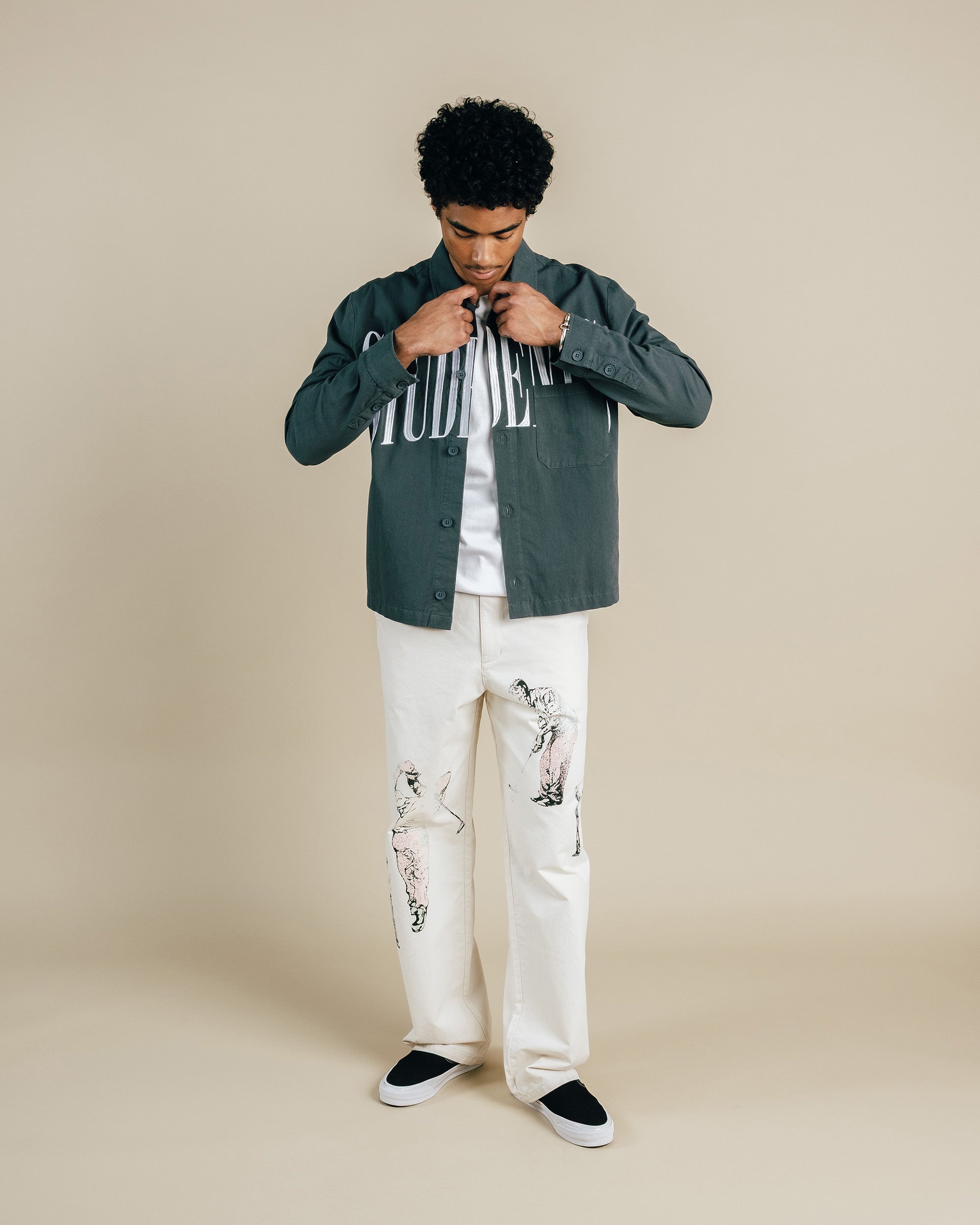 Man wearing a green jacket over a striped shirt with white pants on a beige background