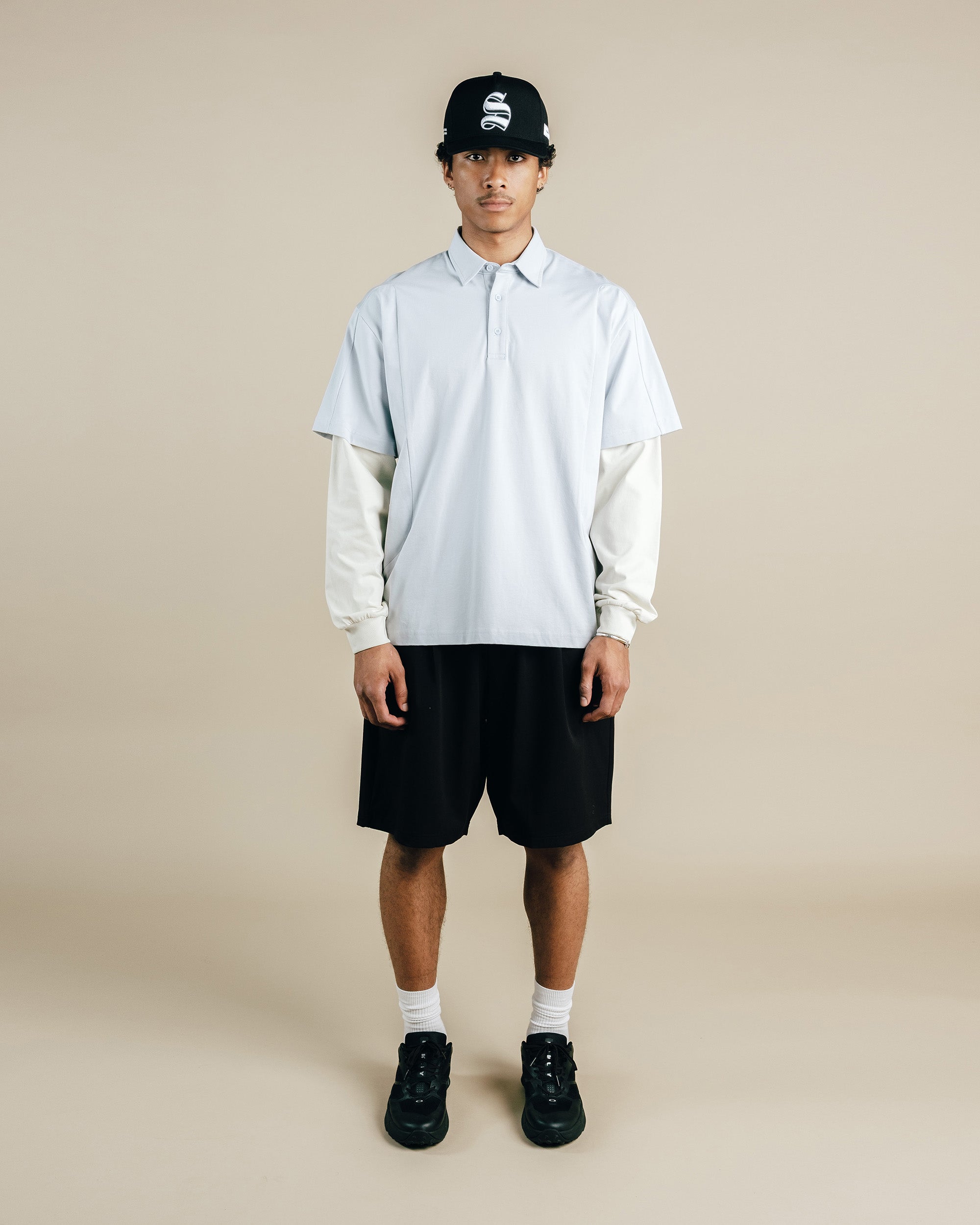 Person wearing a light gray shirt, black shorts, black shoes, and a black cap with a logo on a beige background