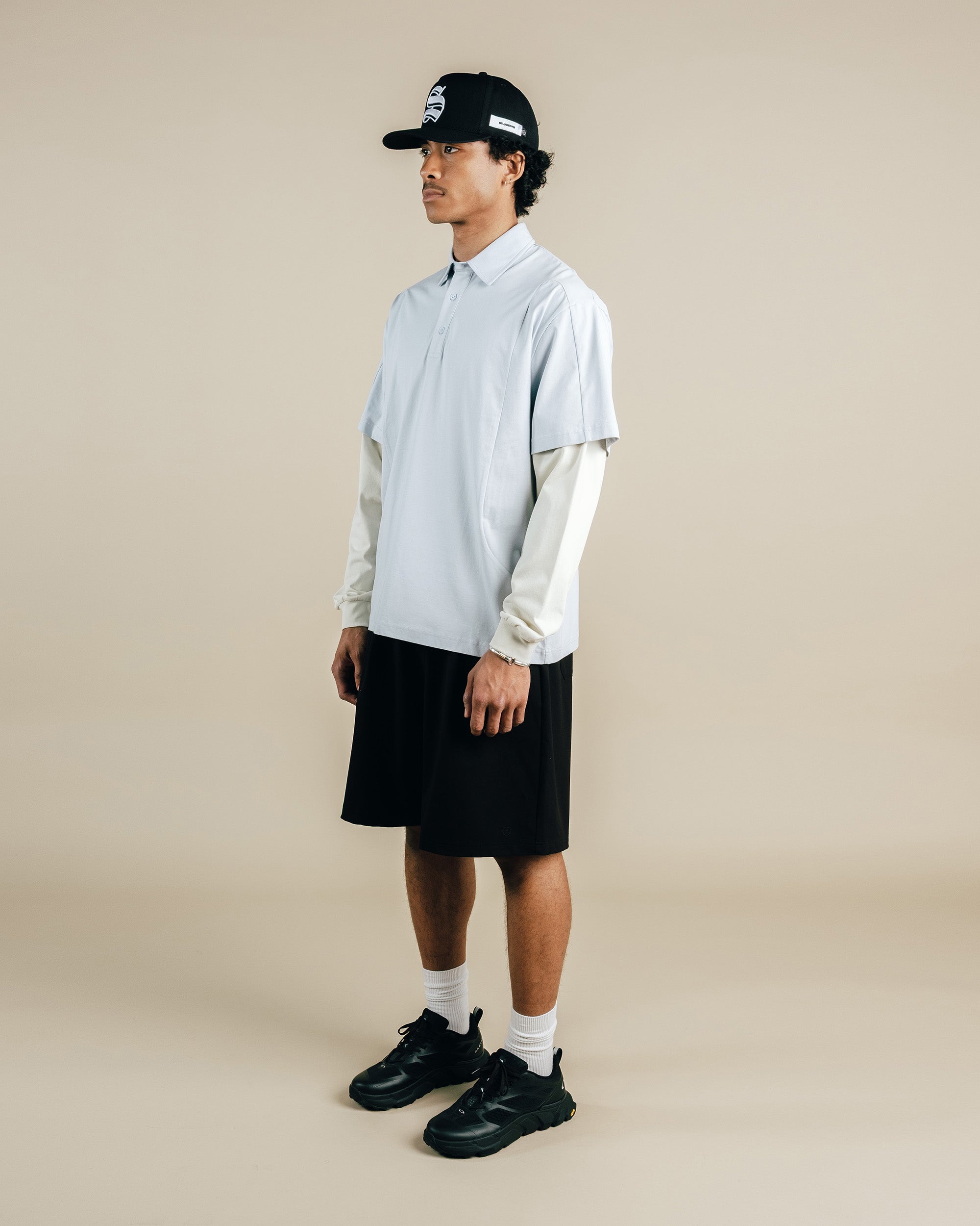 Person wearing a light gray long-sleeve shirt, black shorts, and black sneakers on a beige background