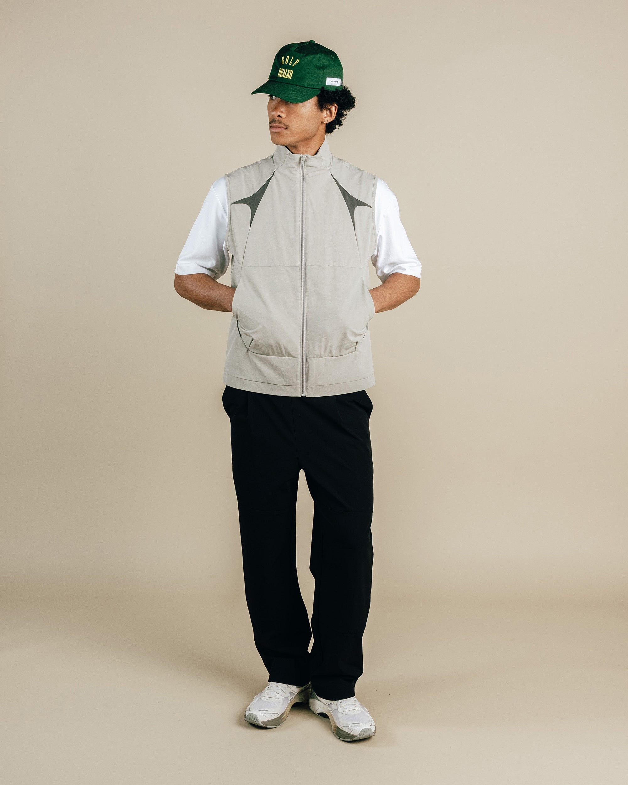 Person wearing a beige vest over a white shirt, black pants, and a green cap on a beige background