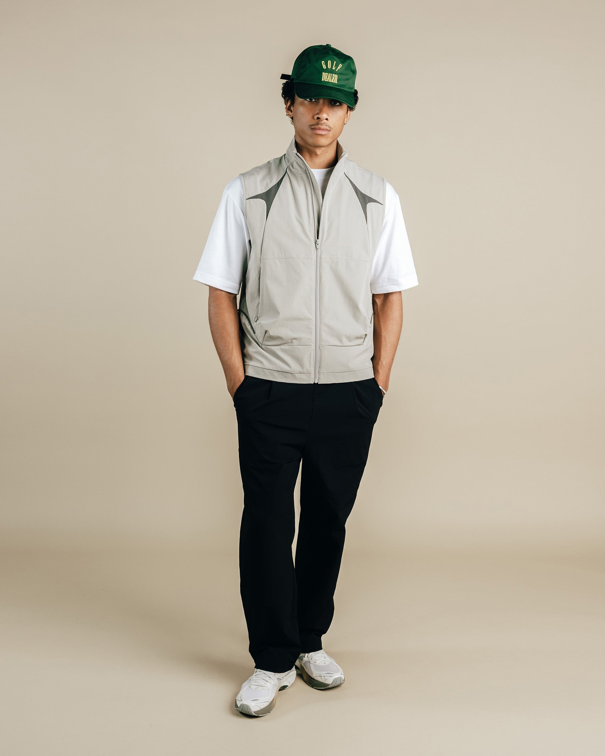 Person wearing a green cap, white shirt, beige vest, black pants, and white sneakers on a beige background