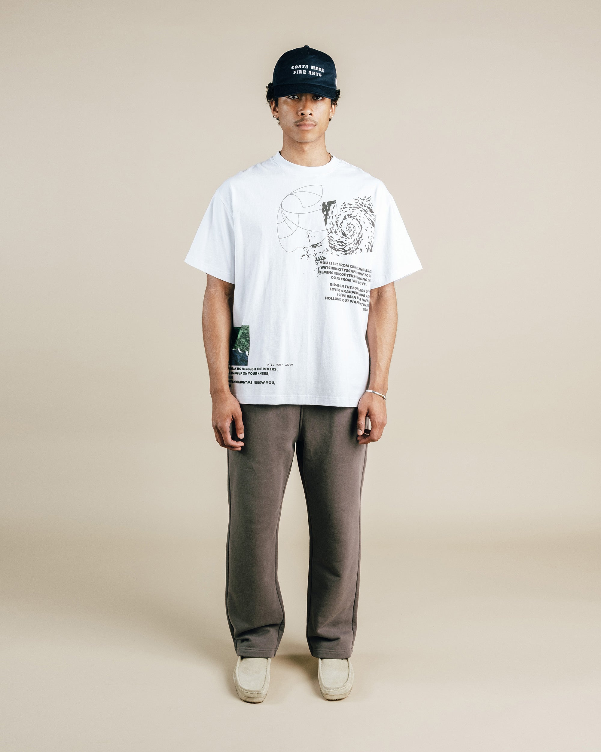 Person wearing a white t-shirt with graphics and text, dark pants, and a cap on a beige background