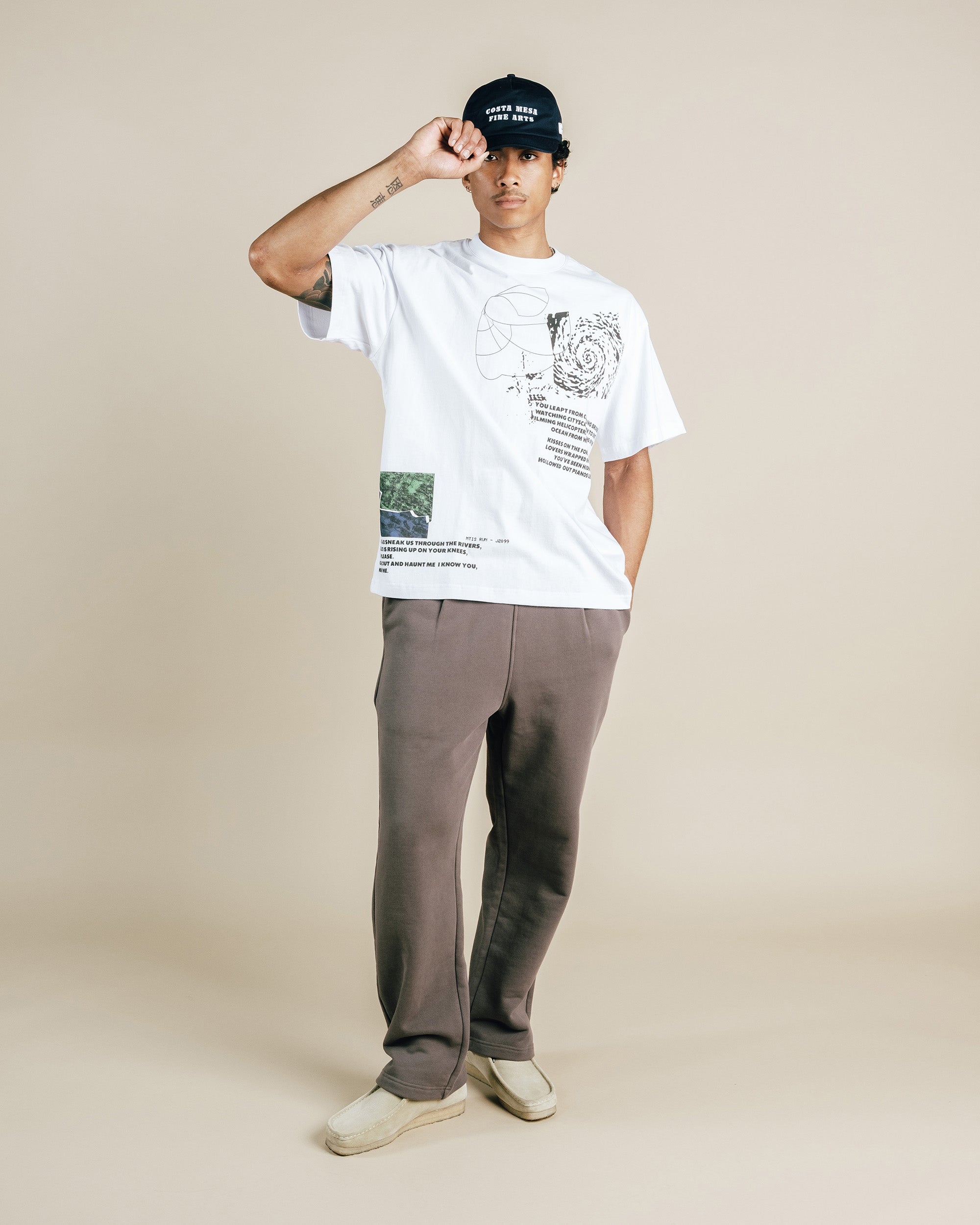 Person wearing a white t-shirt with graphics and text, gray pants, and a black cap on a beige background