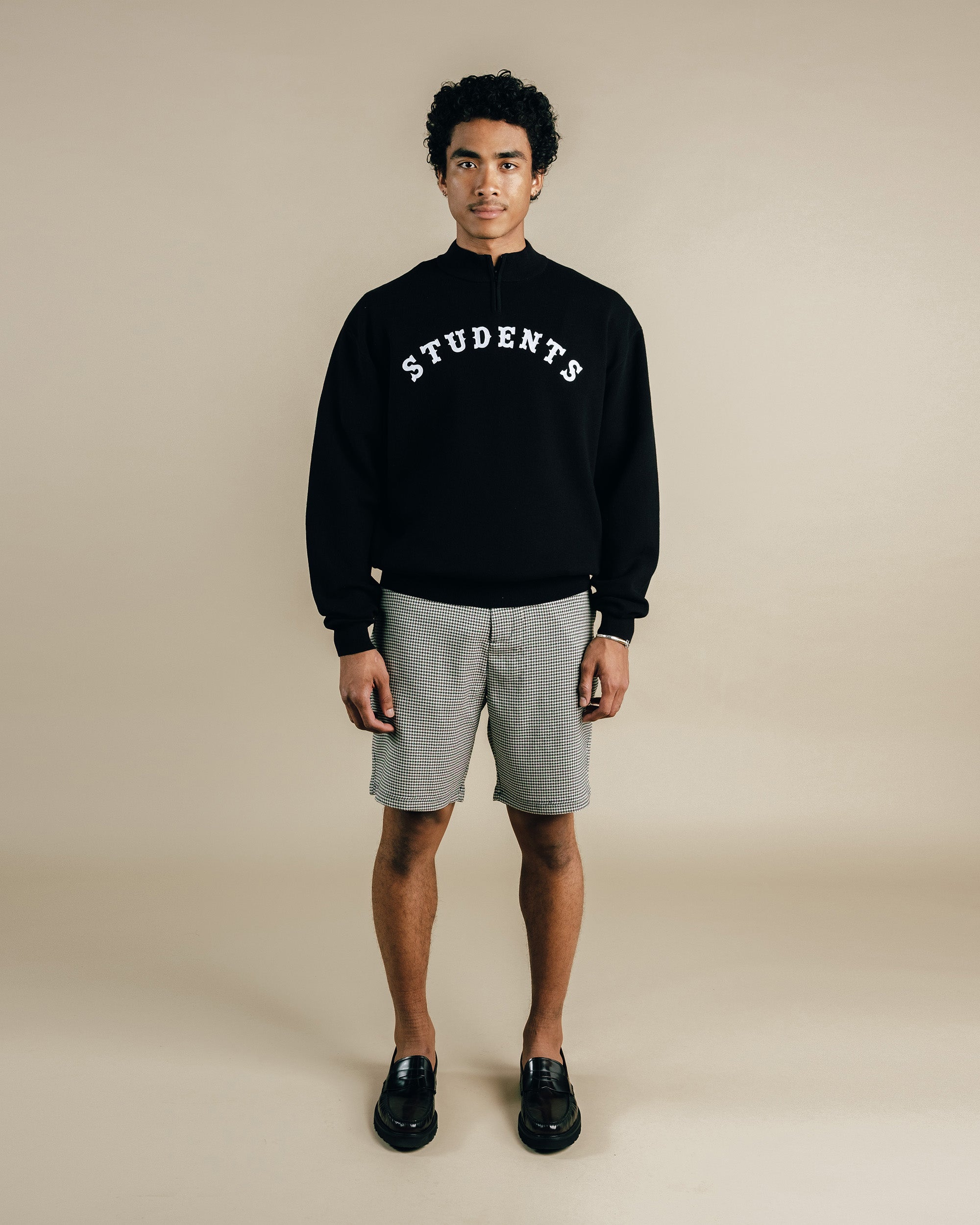 Person wearing a black sweatshirt with 'STUDENTS' text and gray shorts on a beige background