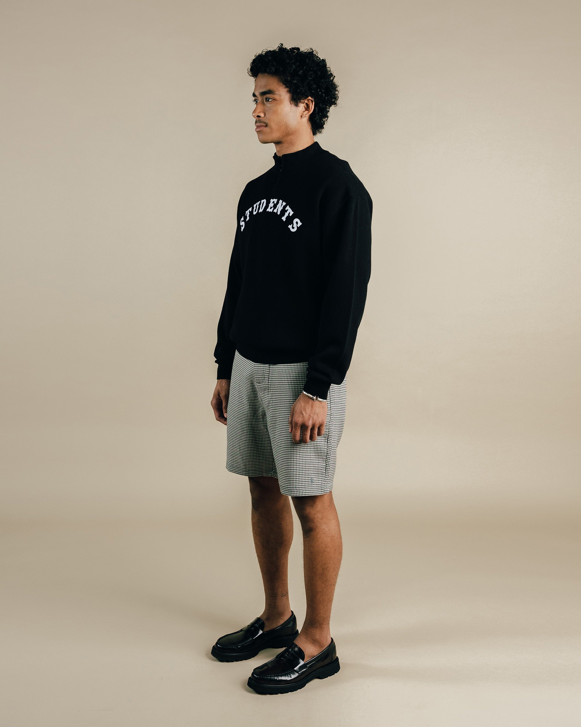 Man wearing a black sweatshirt with text, gray shorts, and black shoes on a beige background