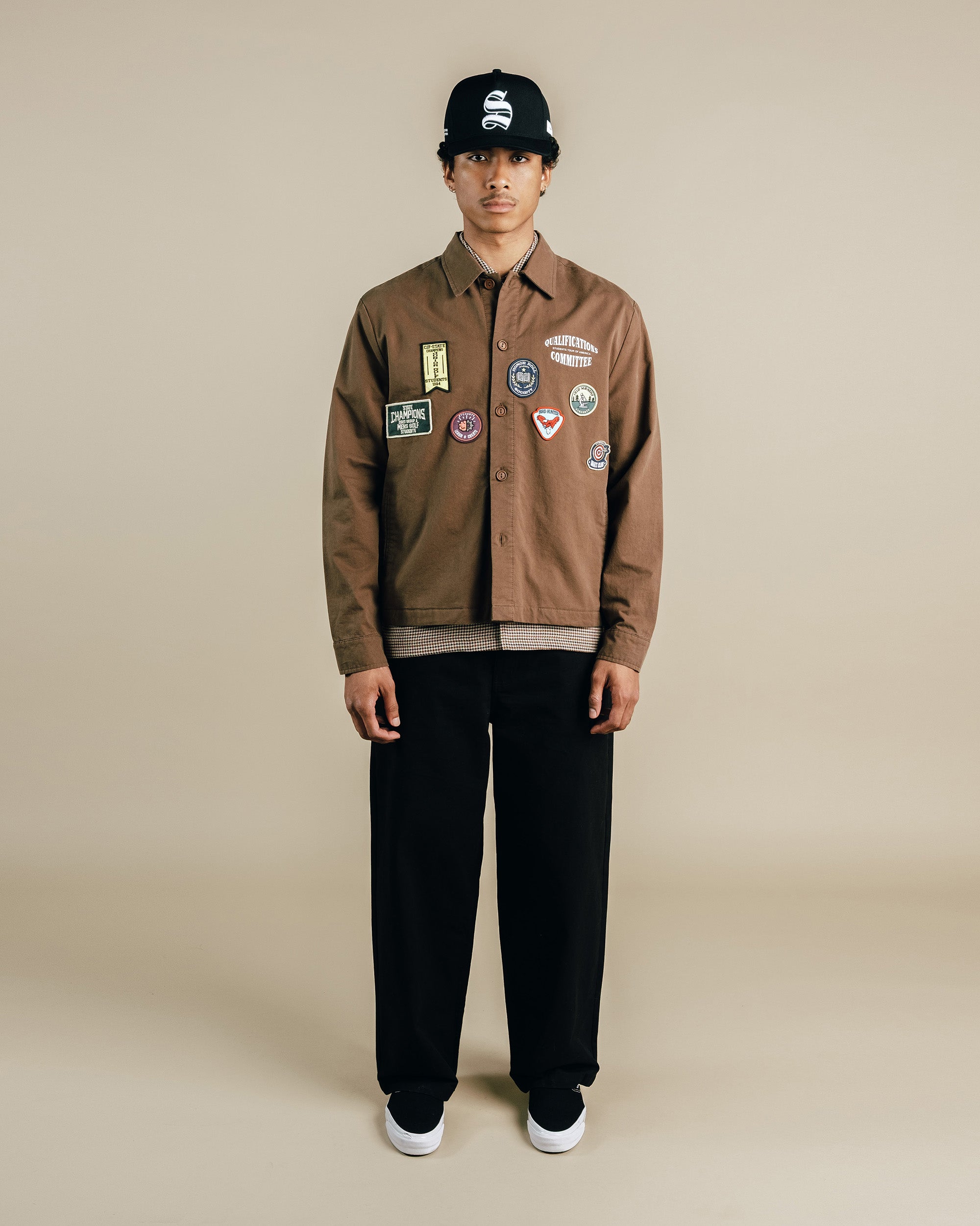 Person wearing a brown jacket with patches, black pants, and a black cap on a beige background