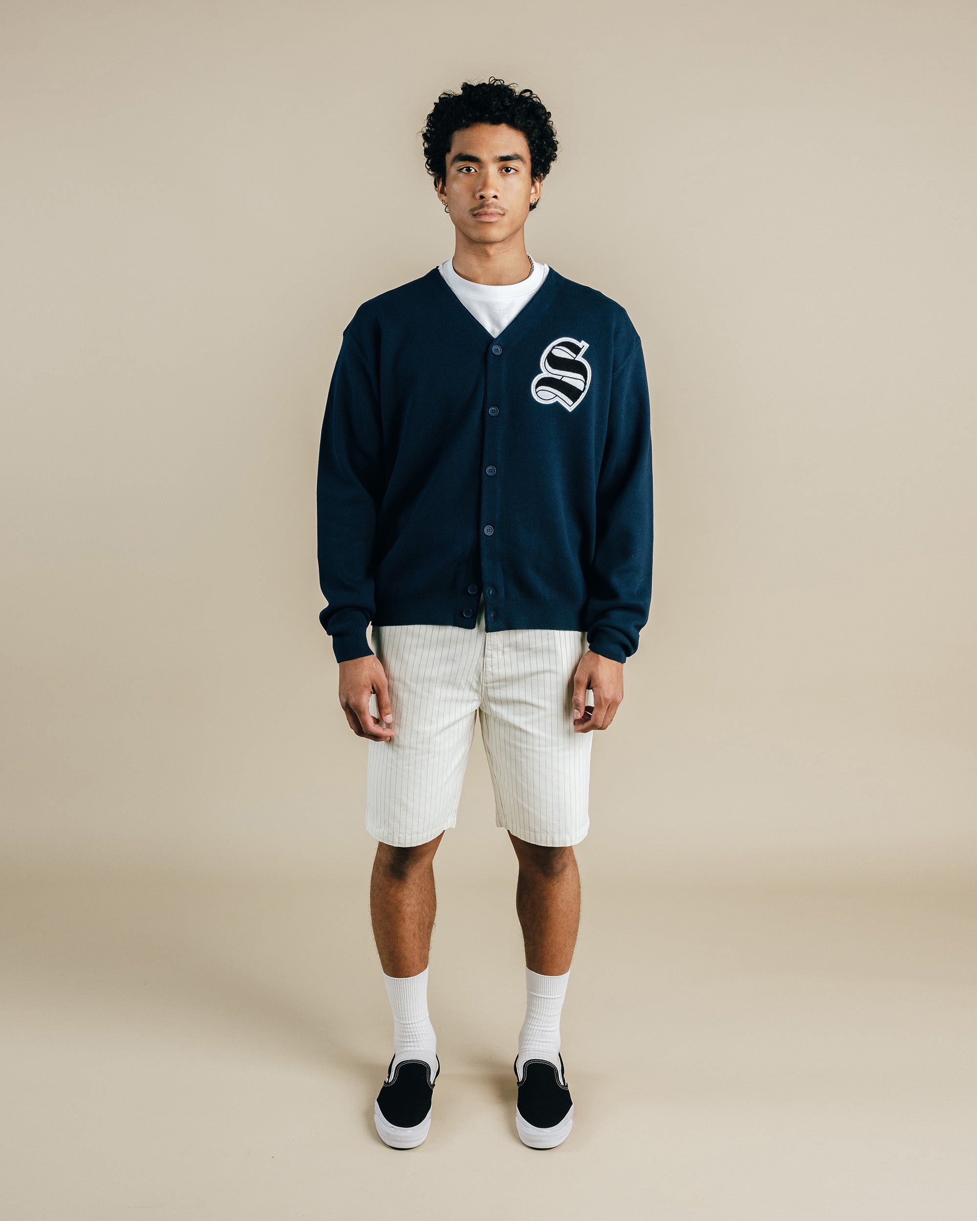 Person wearing a navy cardigan with a logo, white shirt, shorts, and black shoes on a beige background