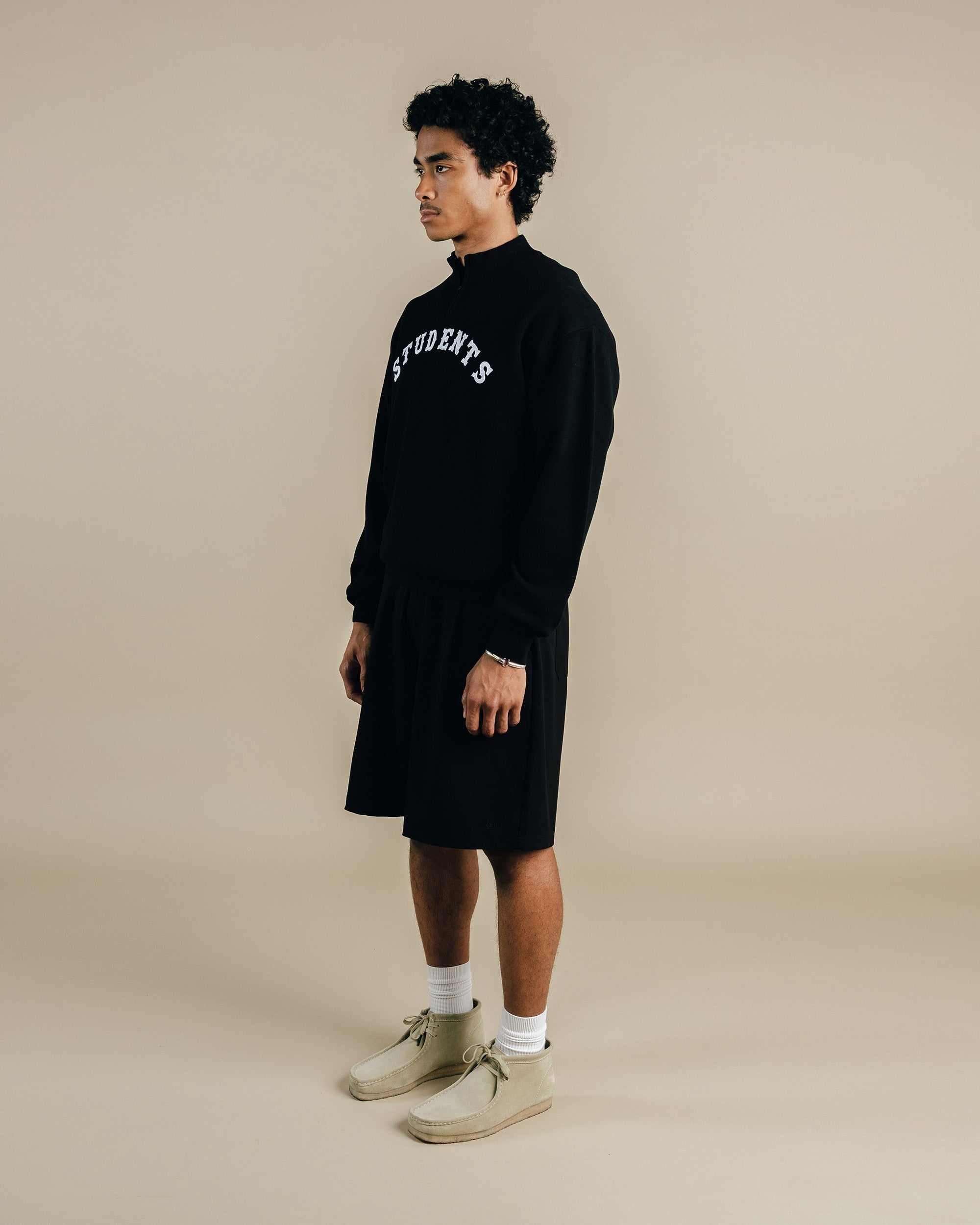 Person wearing a black sweatshirt with text, black shorts, white socks, and beige shoes on a beige background