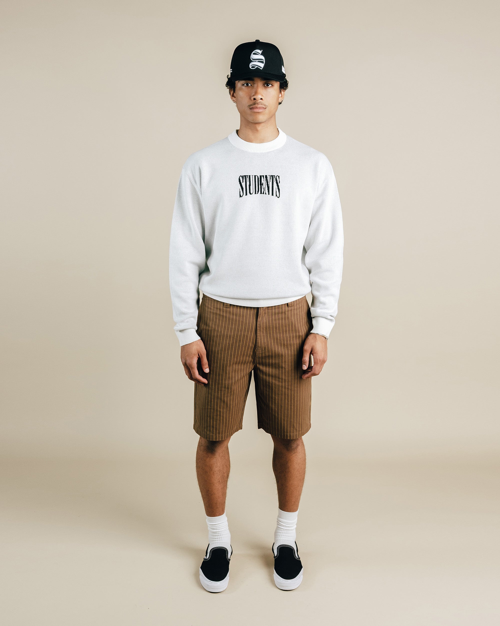 Person wearing a white sweatshirt with 'STUDENTS' text, brown plaid shorts, and black cap on a beige background
