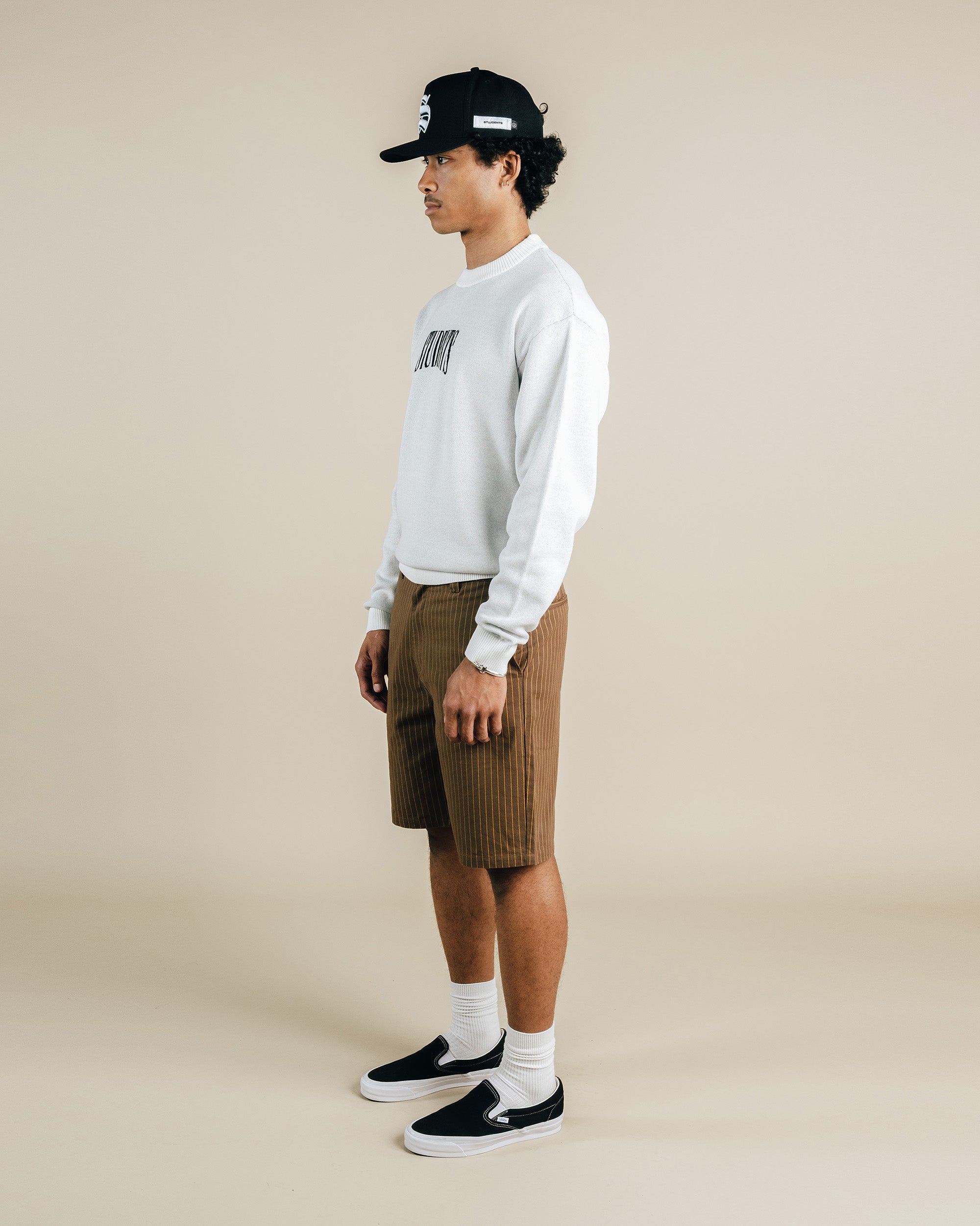 Person wearing a white sweatshirt, brown shorts, and black cap on a beige background