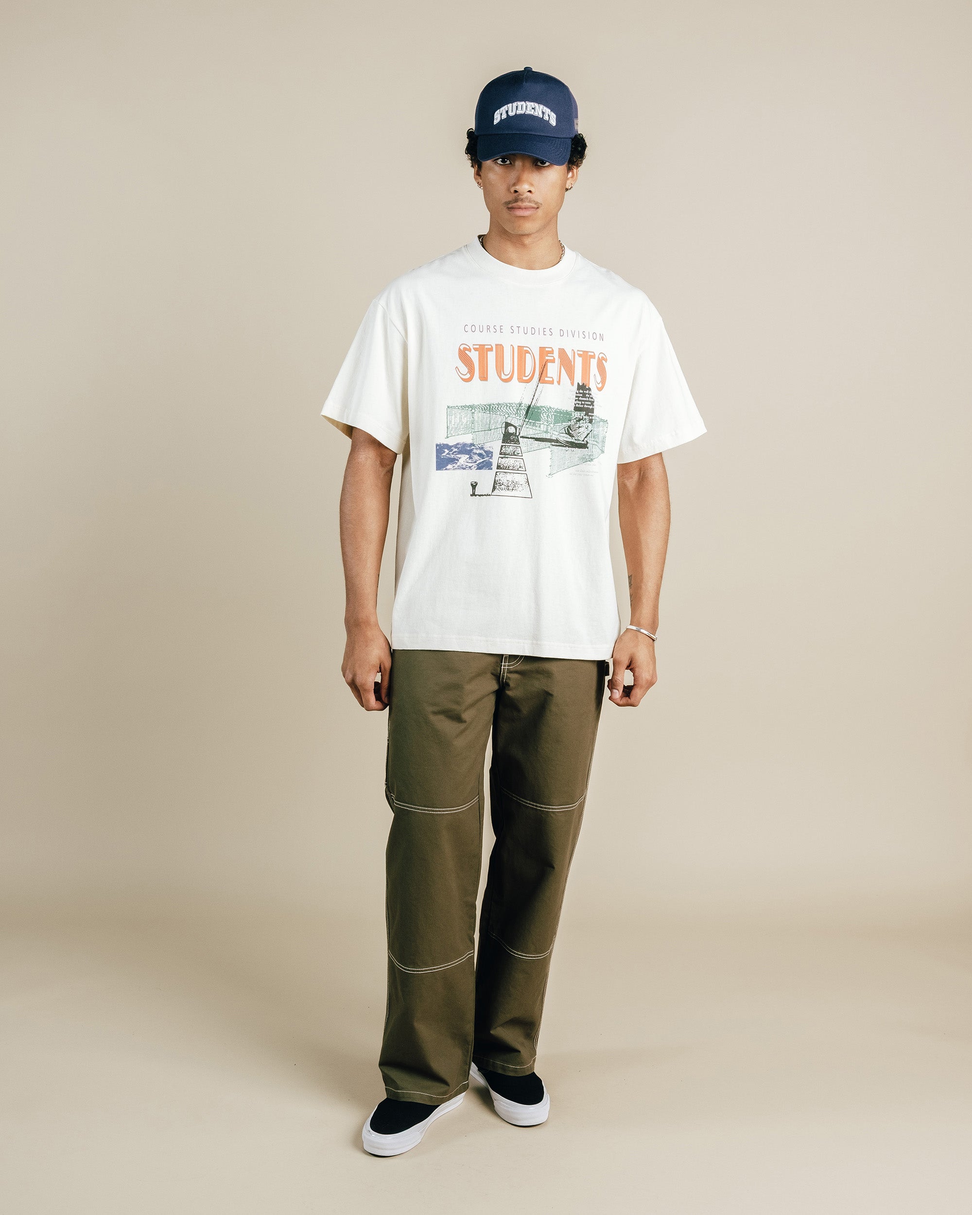 Person wearing a nude t-shirt with graphics and text, green pants, and a navy cap on a beige background