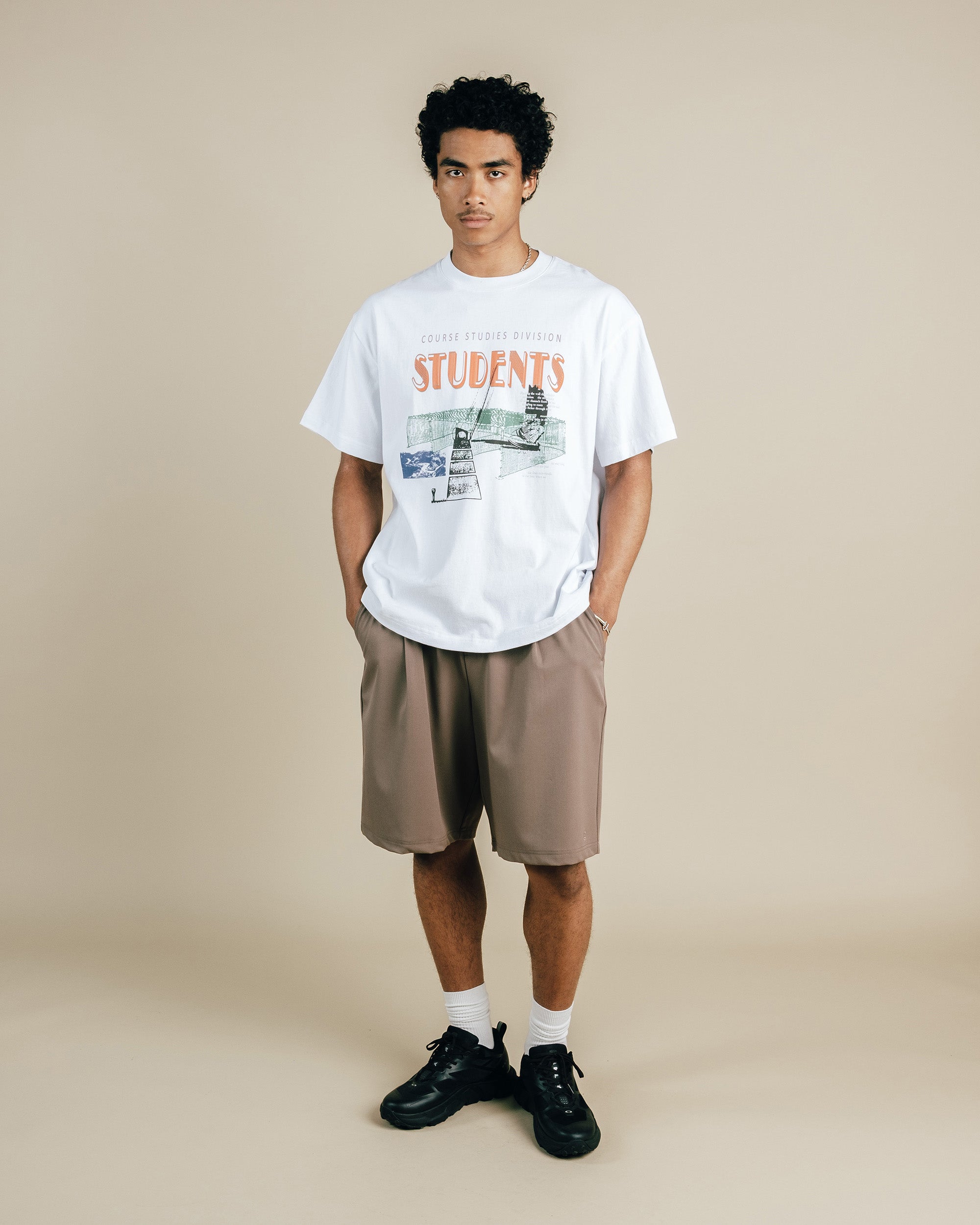 Person wearing a white t-shirt with graphics and text, beige shorts, and black shoes on a beige background