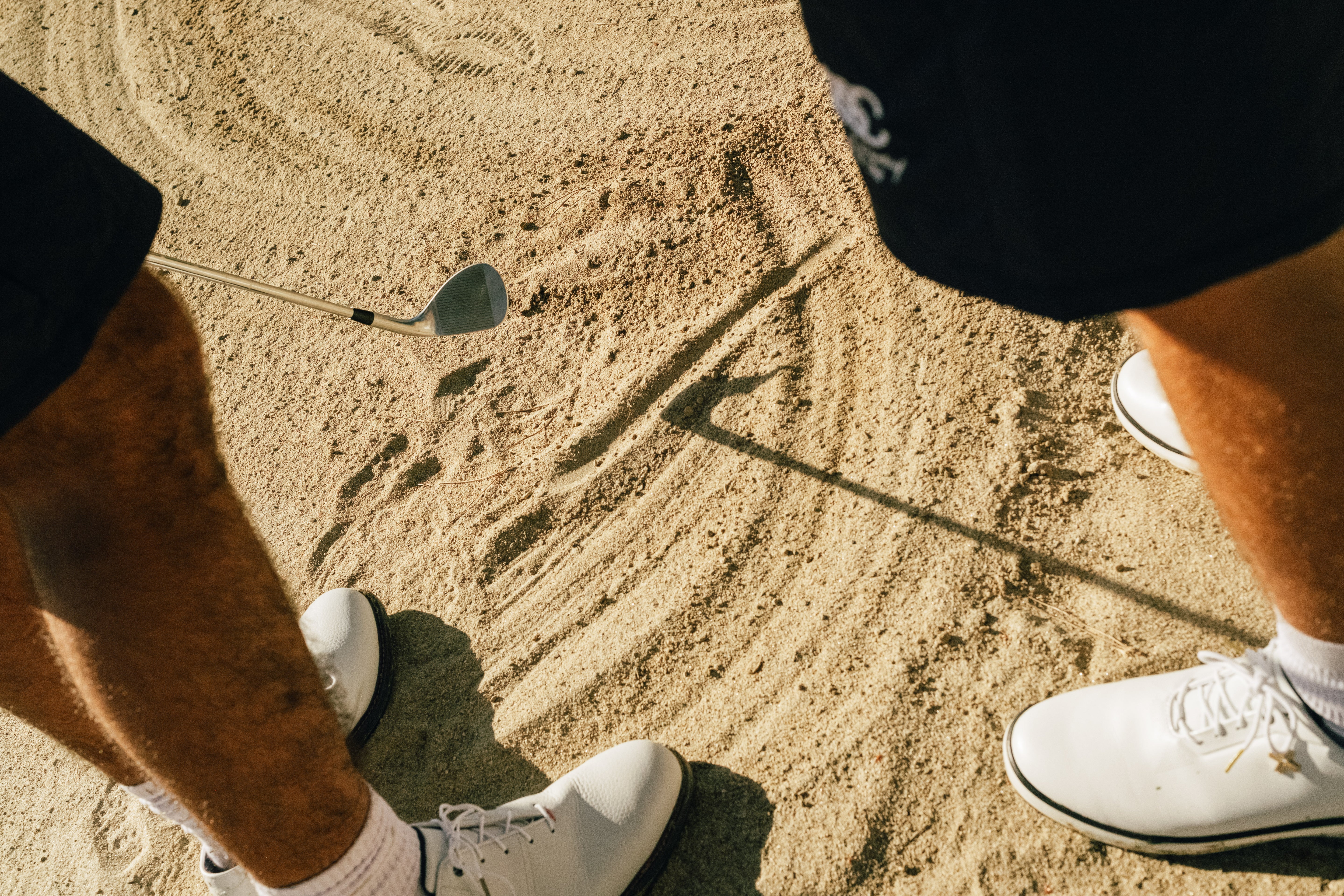 Person in white golf shoes standing on sand with a golf club embedded in the ground.