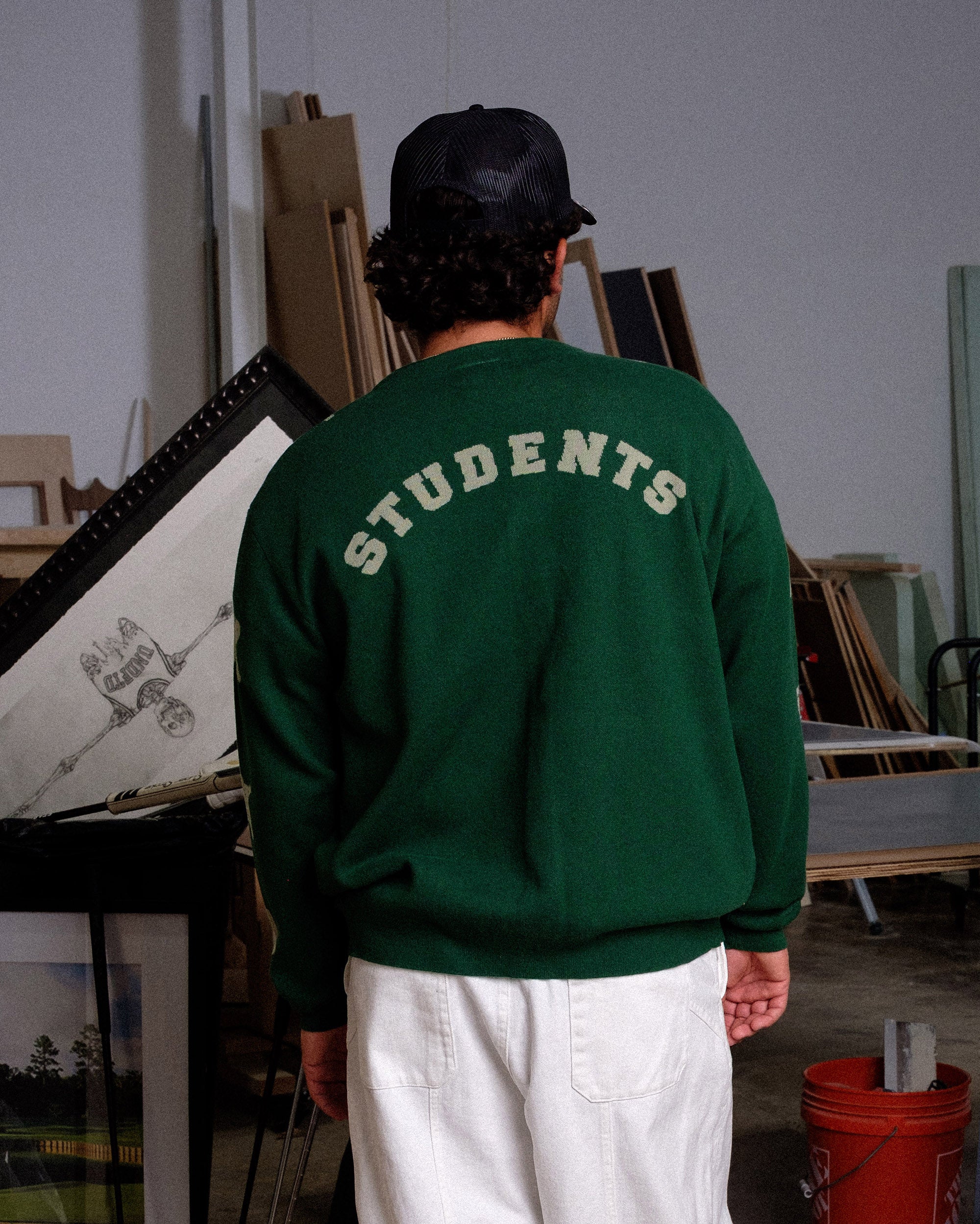 Person wearing a green sweatshirt with 'STUDENTS' text, standing in an indoor setting with artworks.