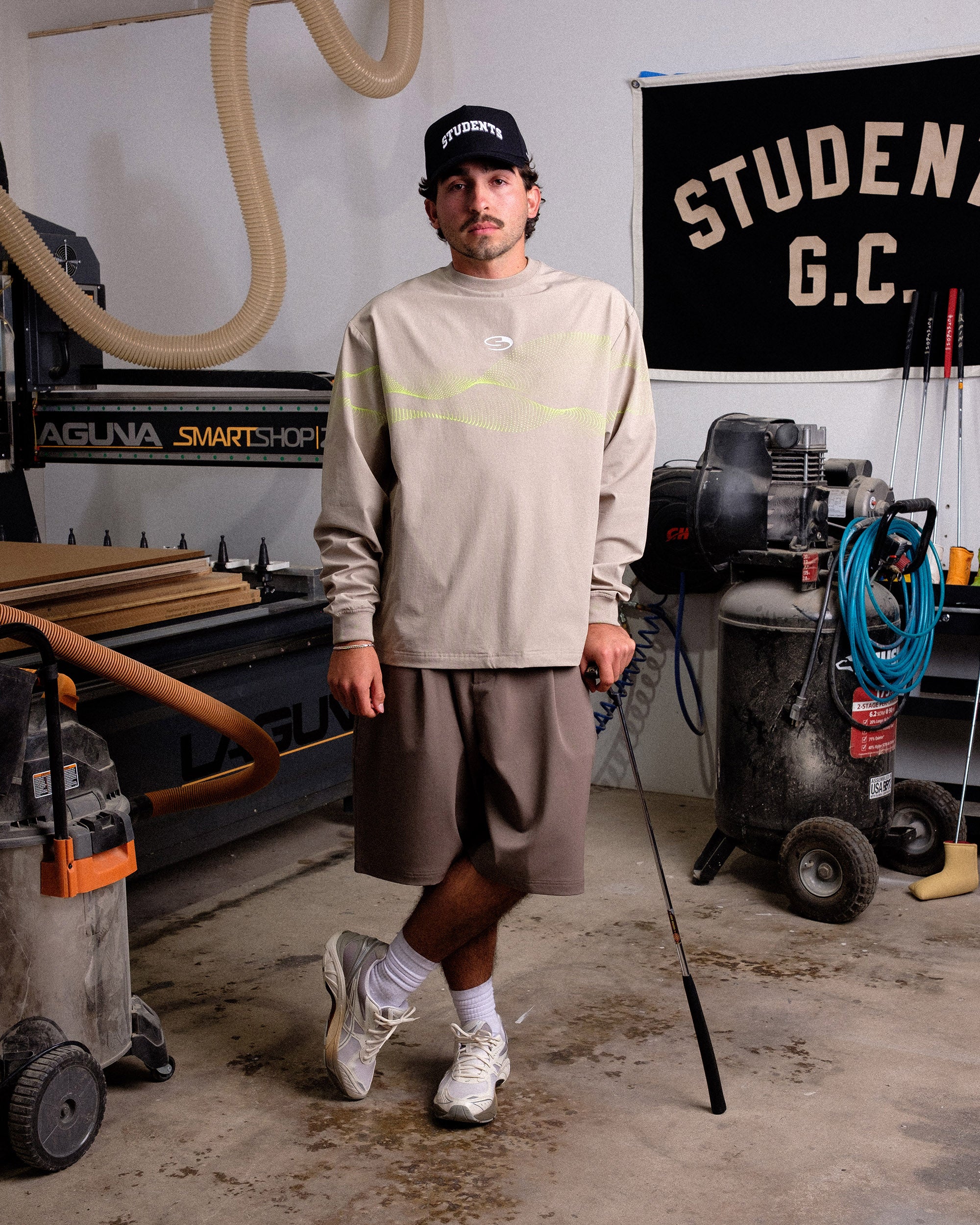 Man standing in a workshop wearing a beige long-sleeve shirt and brown shorts.