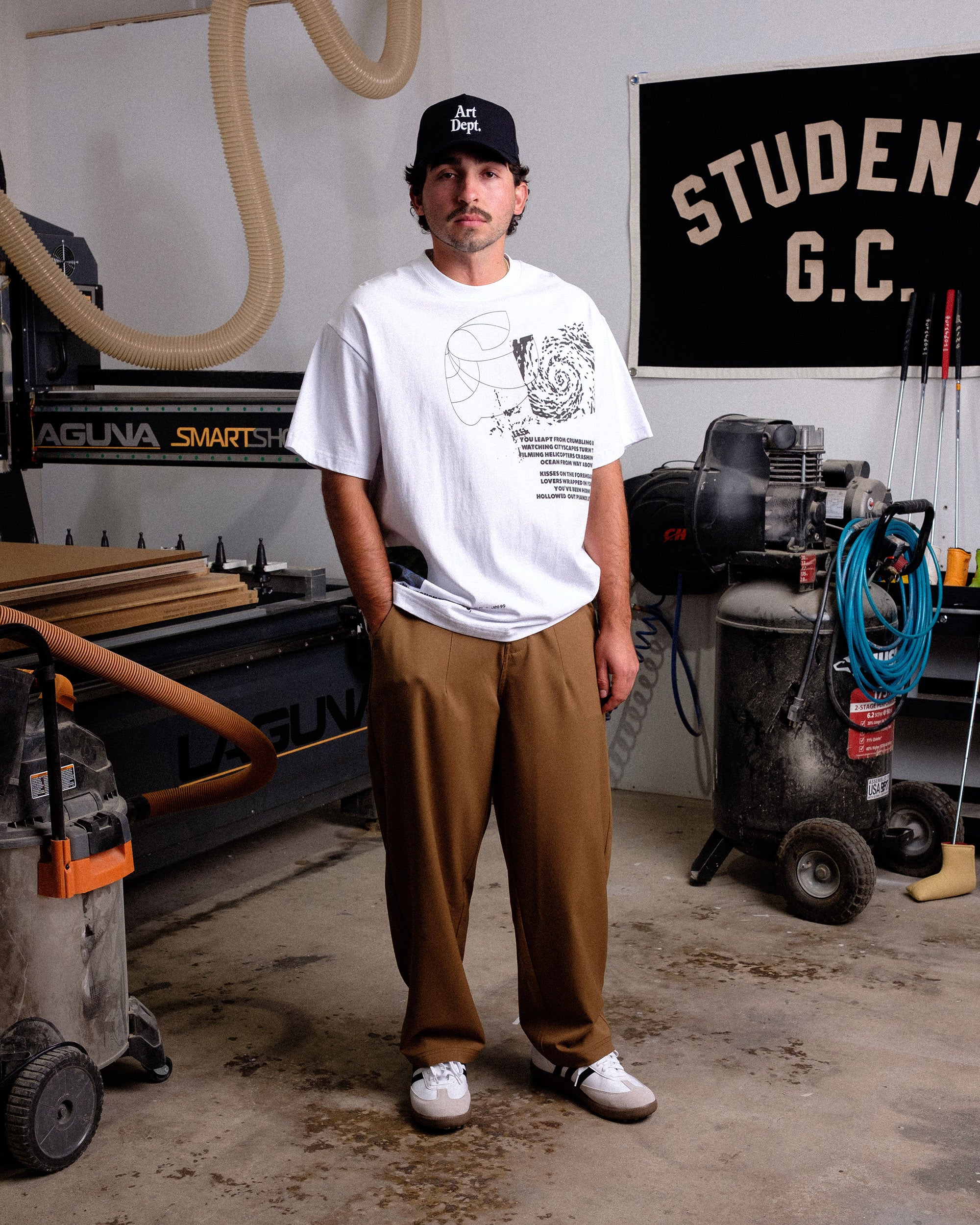 Man standing in a workshop wearing a white t-shirt and brown pants.