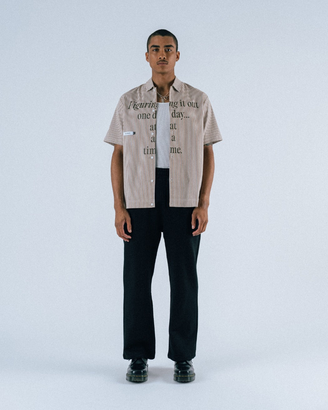 Man wearing a beige shirt with text and black pants on a white background