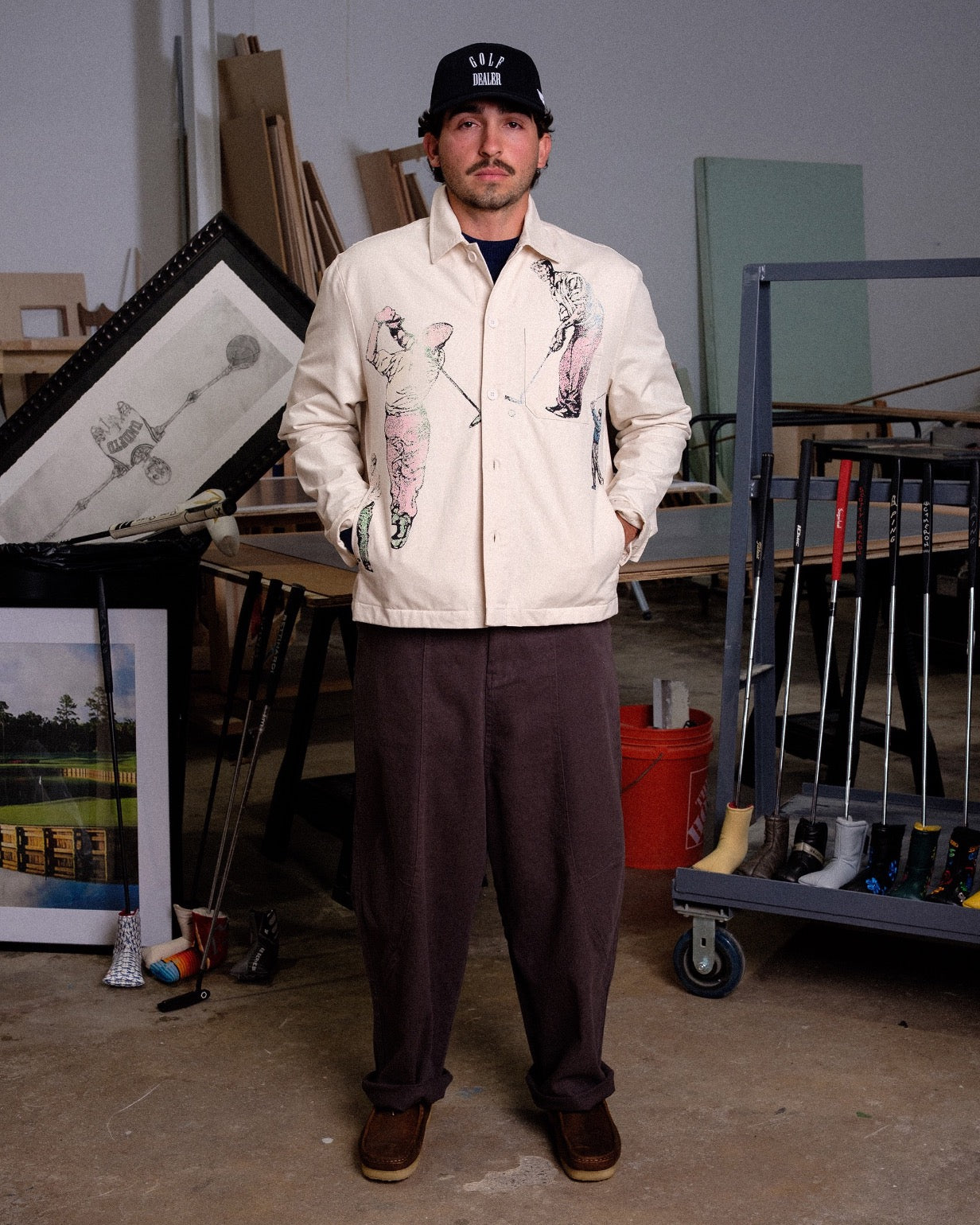 Man wearing a cream jacket with graphic designs in an indoor setting with various items and furniture.