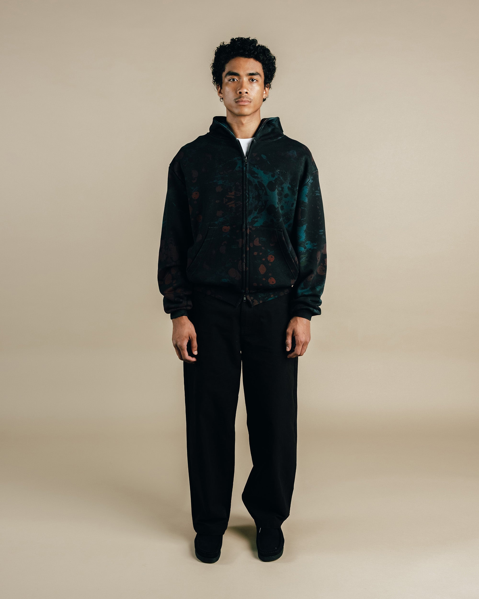 Person wearing a dark patterned jacket and black pants on a beige background