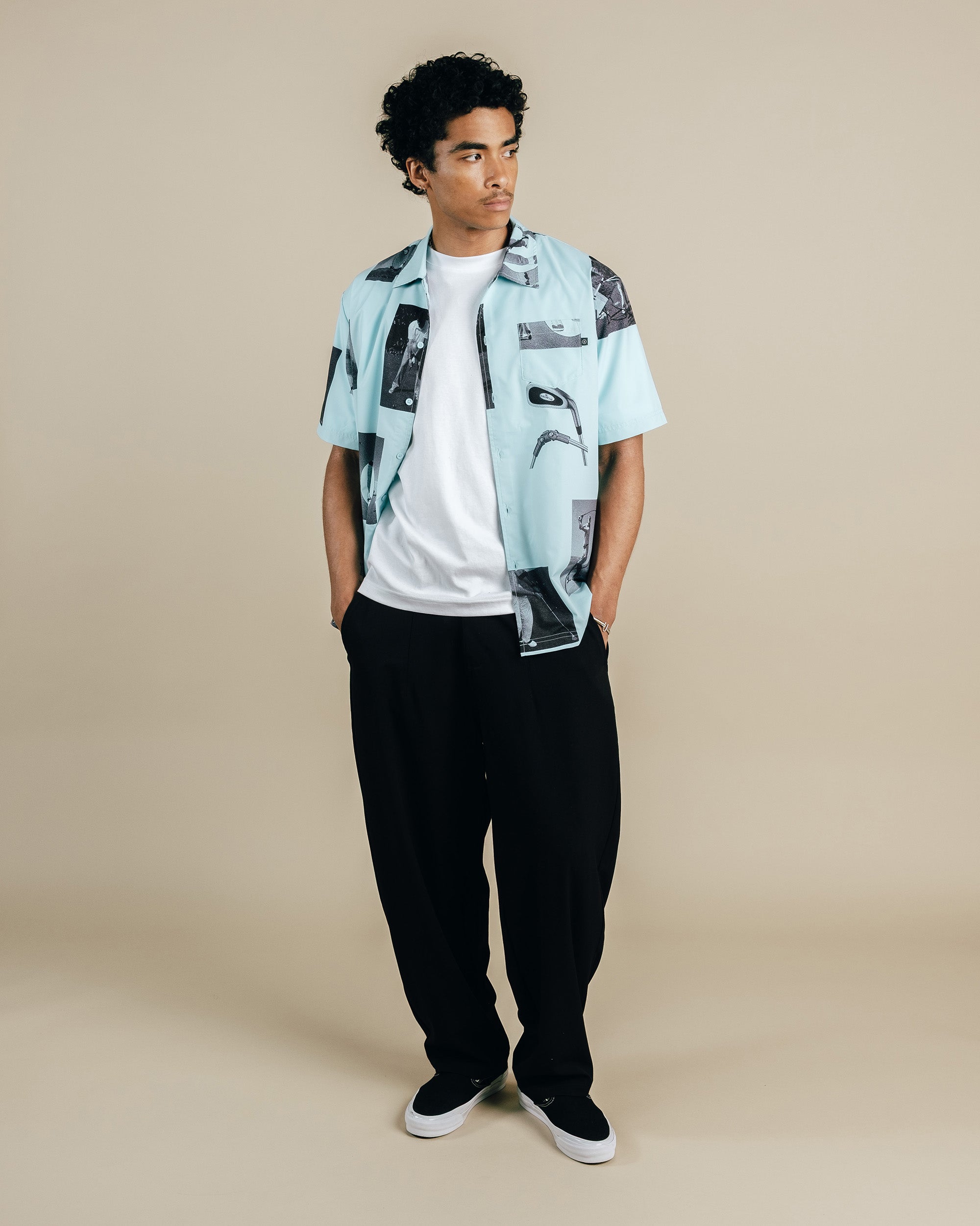 Man wearing a light blue patterned shirt over a white t-shirt with black pants on a beige background