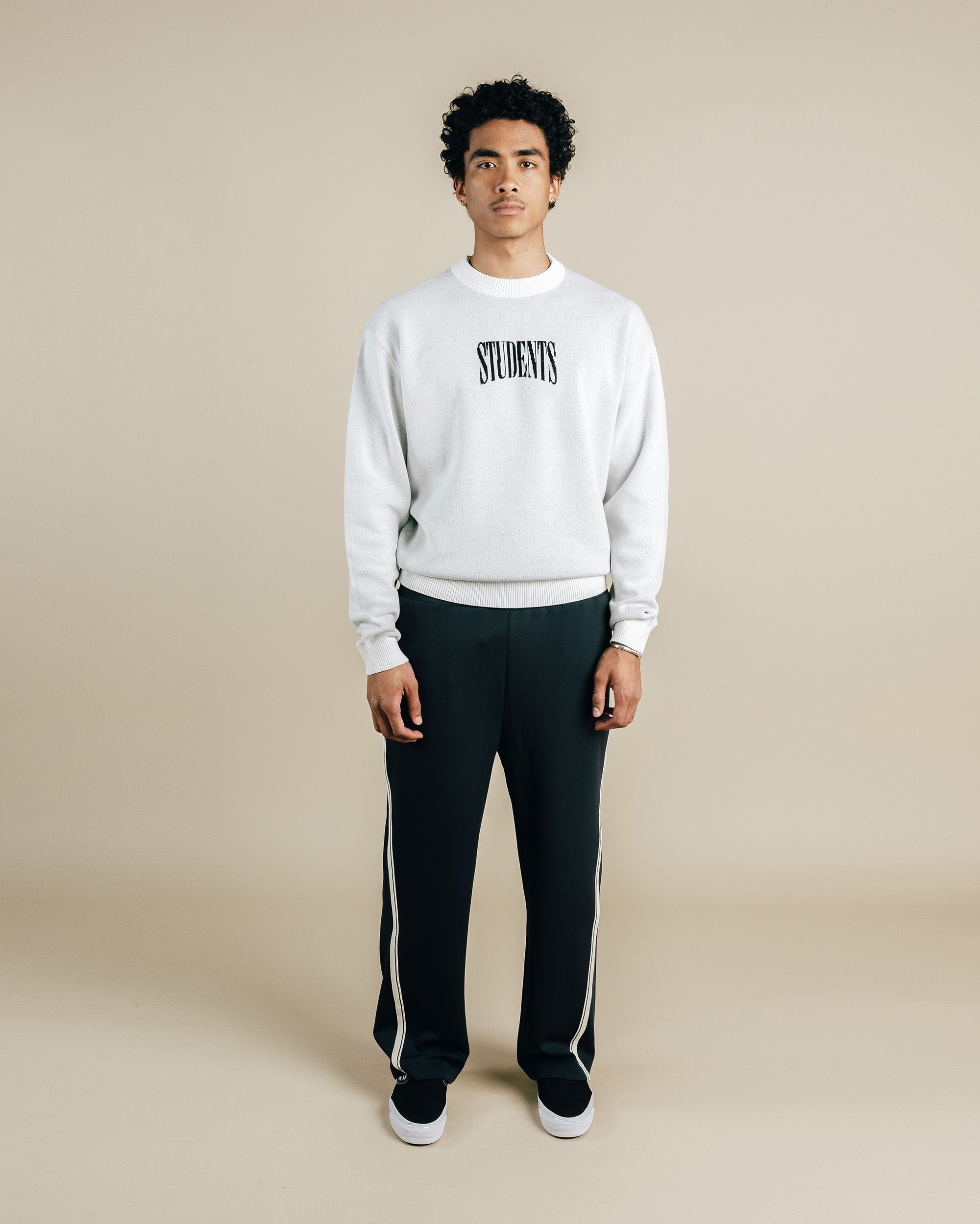 Person wearing a white sweatshirt with 'STUDENTS' text and black pants with white stripes on a beige background